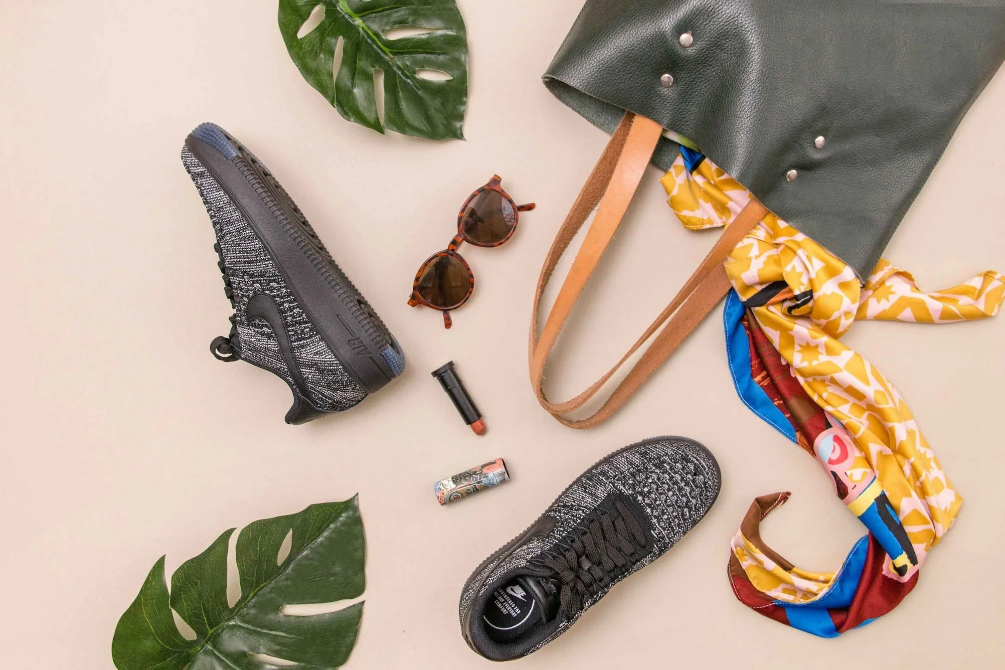 Stylish sneakers, sunglasses, a colorful scarf, an open lipstick, and a purse handle laid out on a peach background.