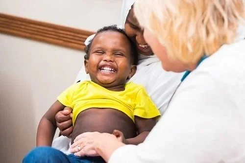 A young, laughing child being held by a parent and getting her stomach checked by a doctor.