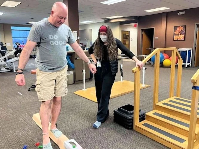 A patient and physical therapist working together, as the patient balances a leg on a wooden beam.