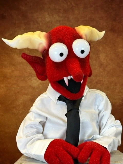A puppet character with red skin, yellow horns, large googly eyes, and fanged mouth, dressed in a white shirt and black tie, against a brown background.