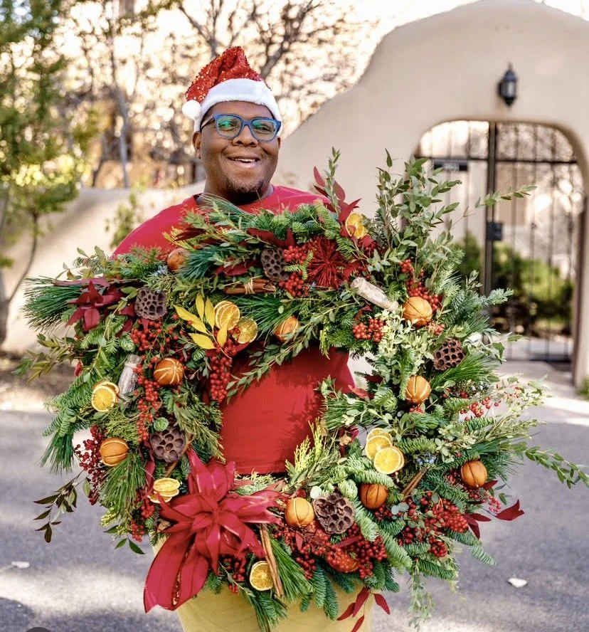 A man wearing a Santa hat and glasses stands outdoors holding a large holiday wreath decorated with oranges, berries, pinecones, and greenery. He is smiling and dressed in a red shirt.