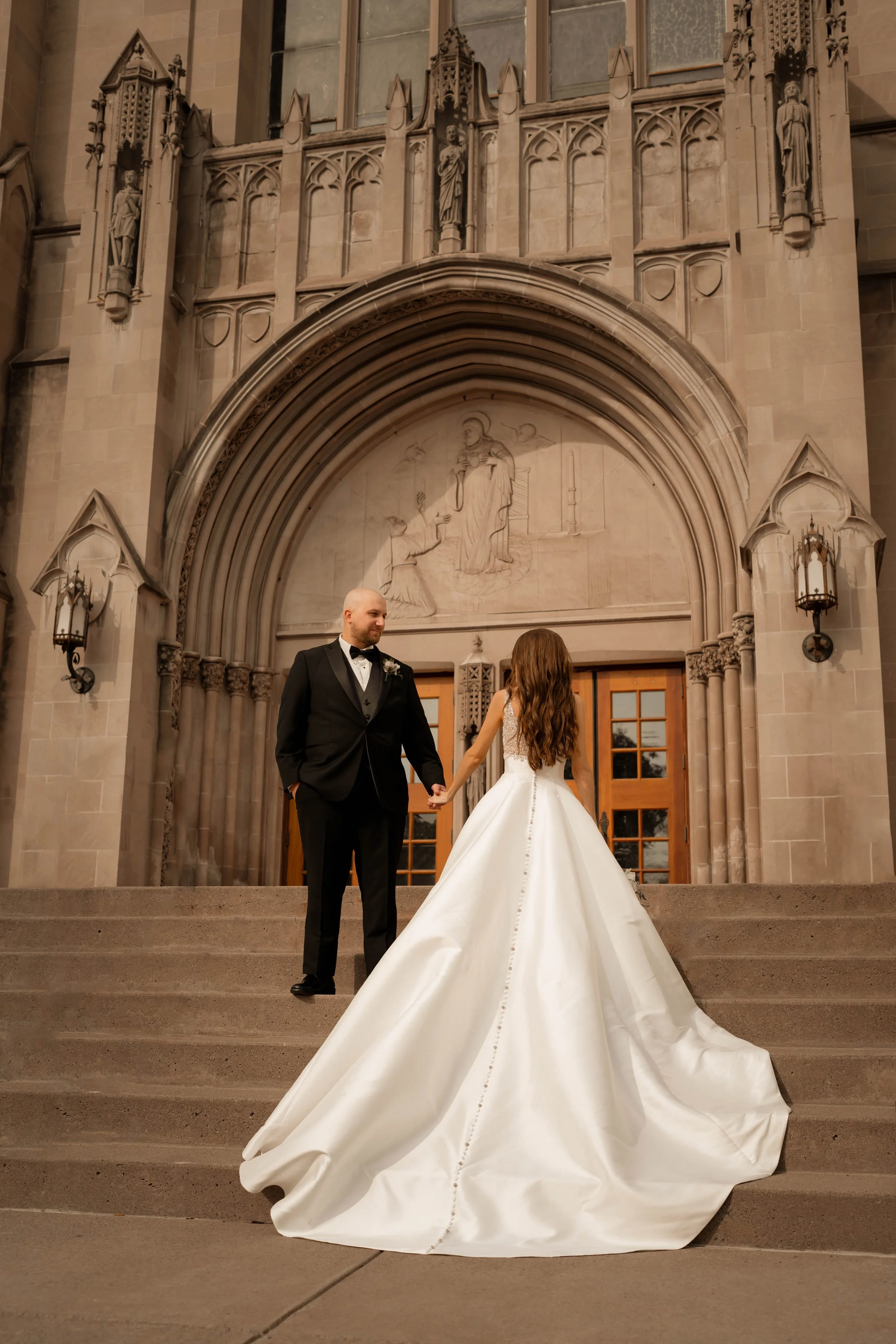 A bride and groom holding hands on the steps outside a church, with the bride in a white wedding gown and the groom in a black tuxedo, at their wedding ceremony.