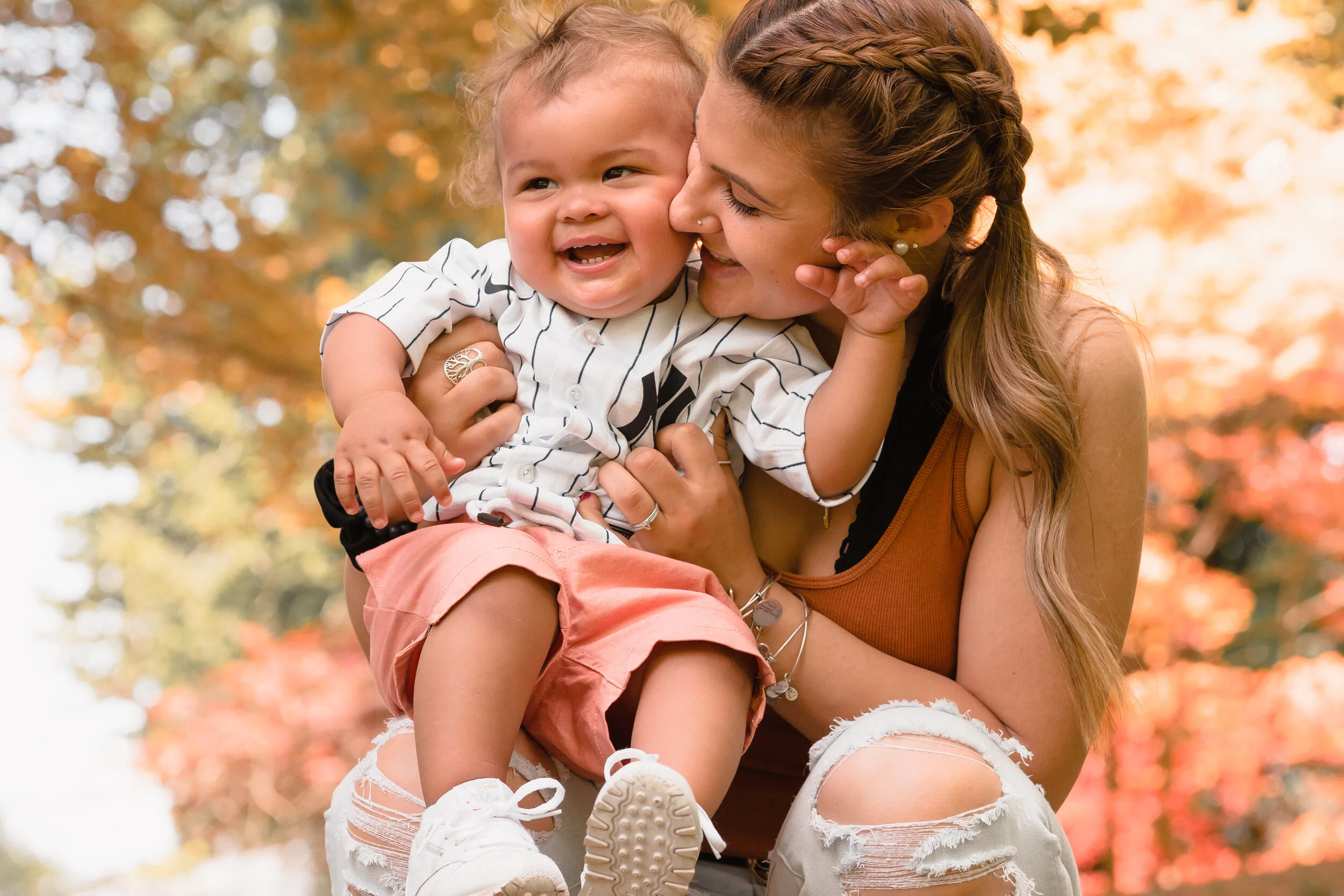 A woman and a young child sharing a joyful moment outdoors amidst fall foliage, with the woman holding the child up in her arms, both smiling and laughing.