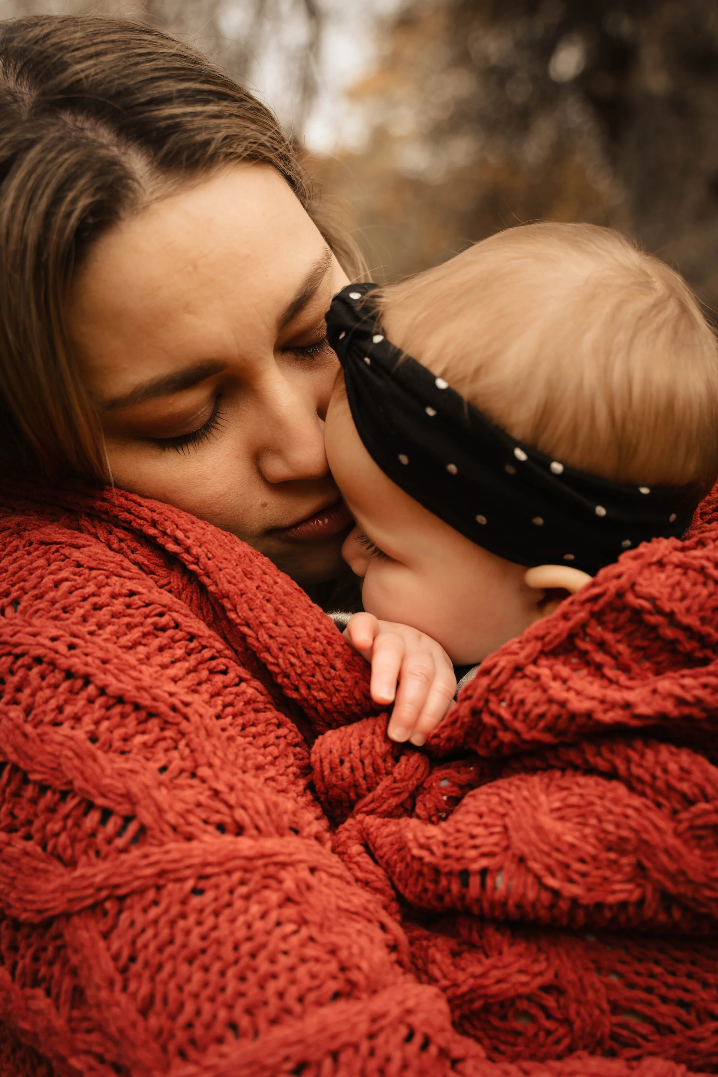 A woman and a baby sharing a gentle, close moment outdoors, both wrapped in red knit blankets, with their faces touching, and the woman holding the baby lovingly.