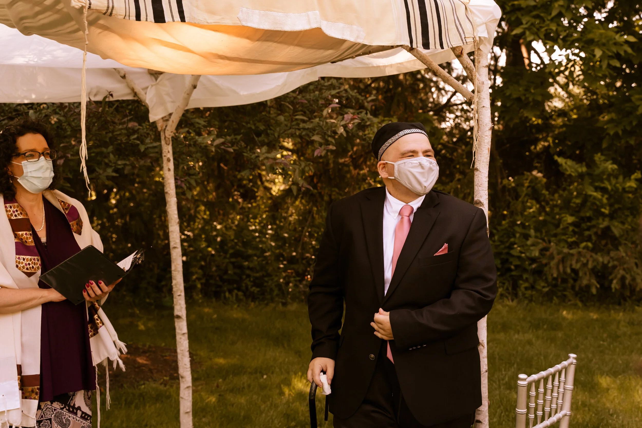 A man in a black suit, pink tie, and face mask standing outdoors under a white canopy with a woman in a face mask holding a book or folder nearby.