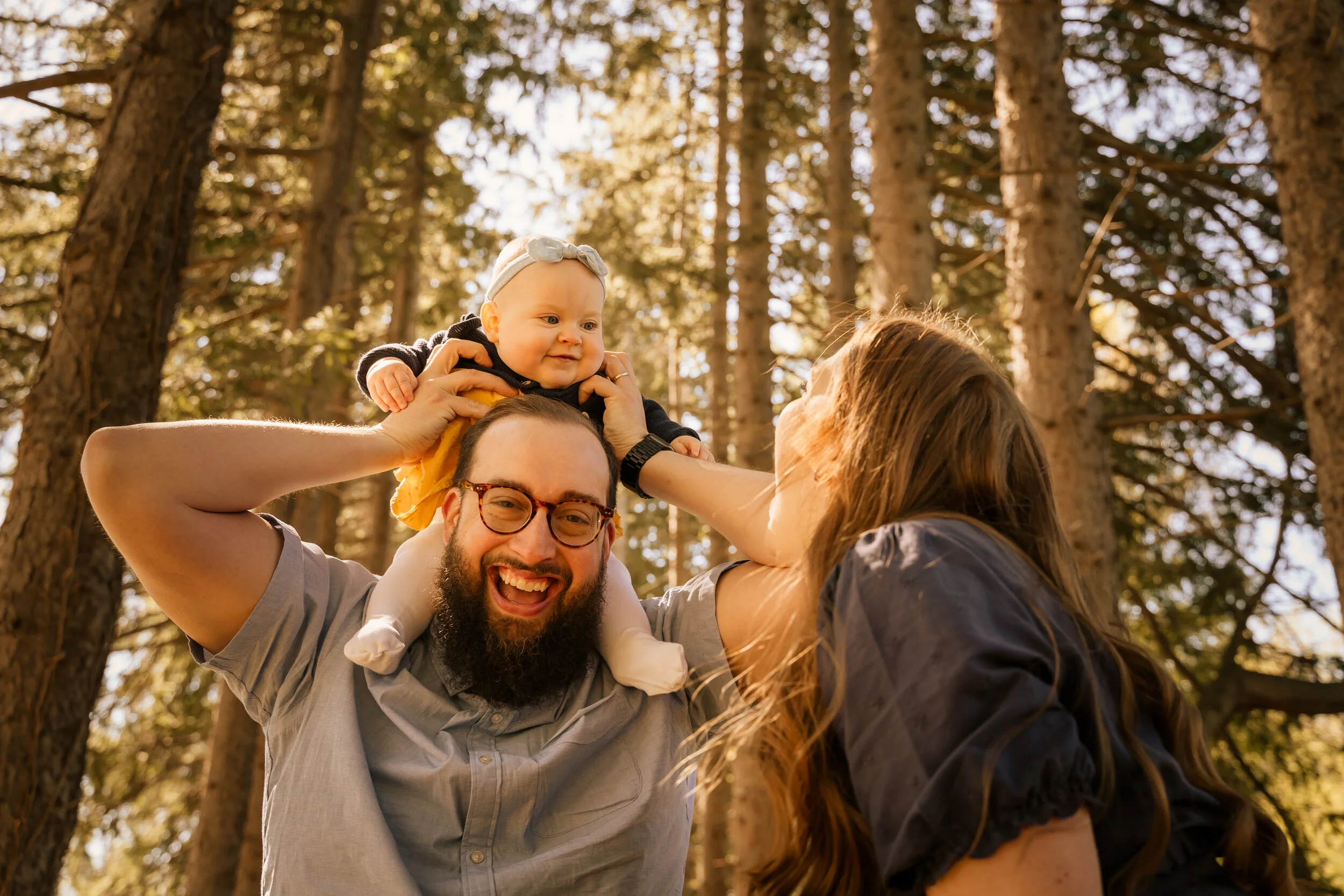A family of three enjoying a sunny day in the woods, with the father holding their baby on his shoulders, and the mother reaching up and touching the baby's face.