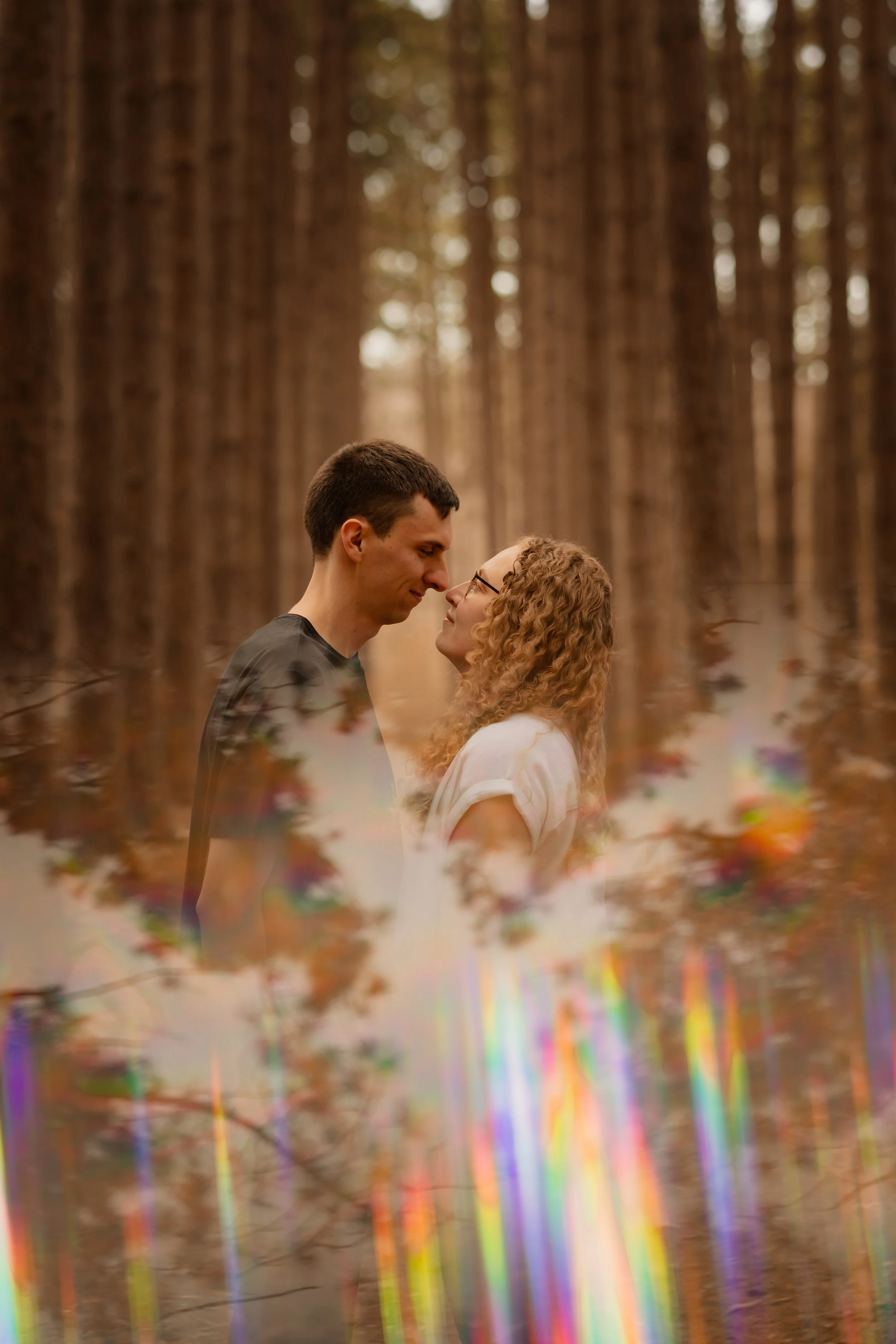 A couple standing close together in a forest, facing each other tenderly with their noses touching. The scene features tall trees in the background and a colorful, abstract overlay at the bottom of the image.