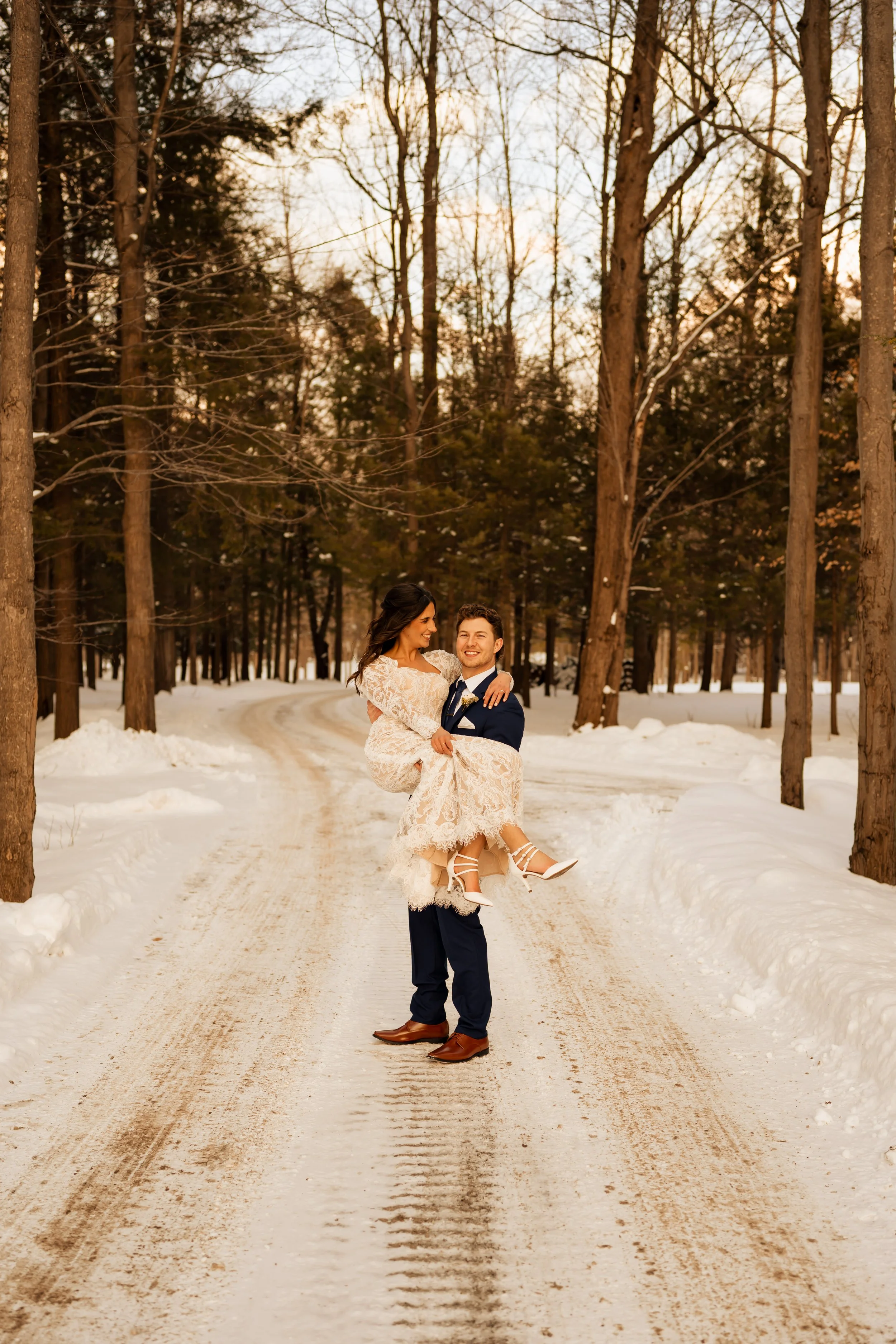 A couple in wedding attire, the groom carrying the bride, standing on a snowy dirt road in a forest during sunset.