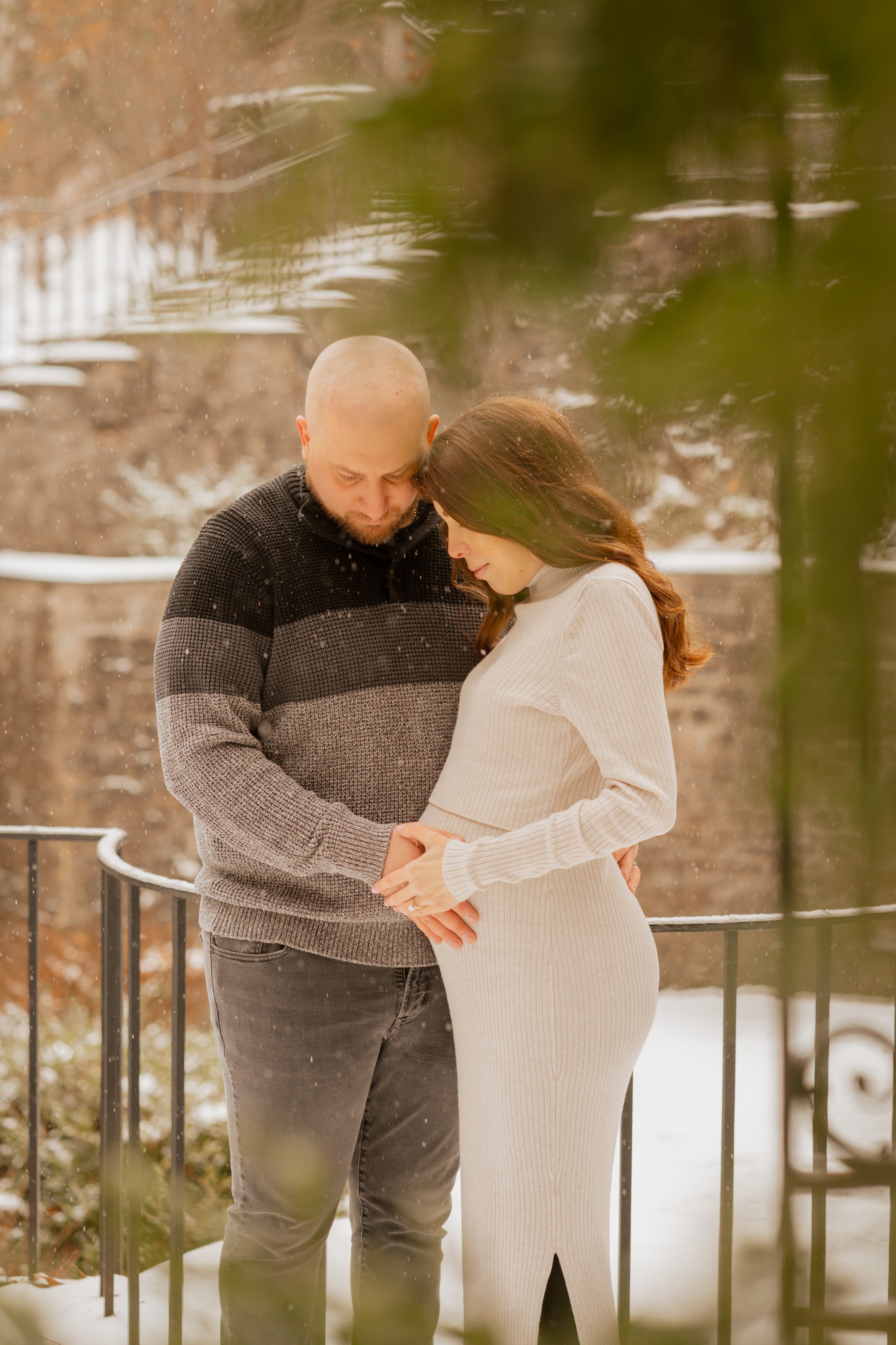 A couple standing outdoors in winter, holding hands and touching foreheads, with snow on the ground and a fence behind them.
