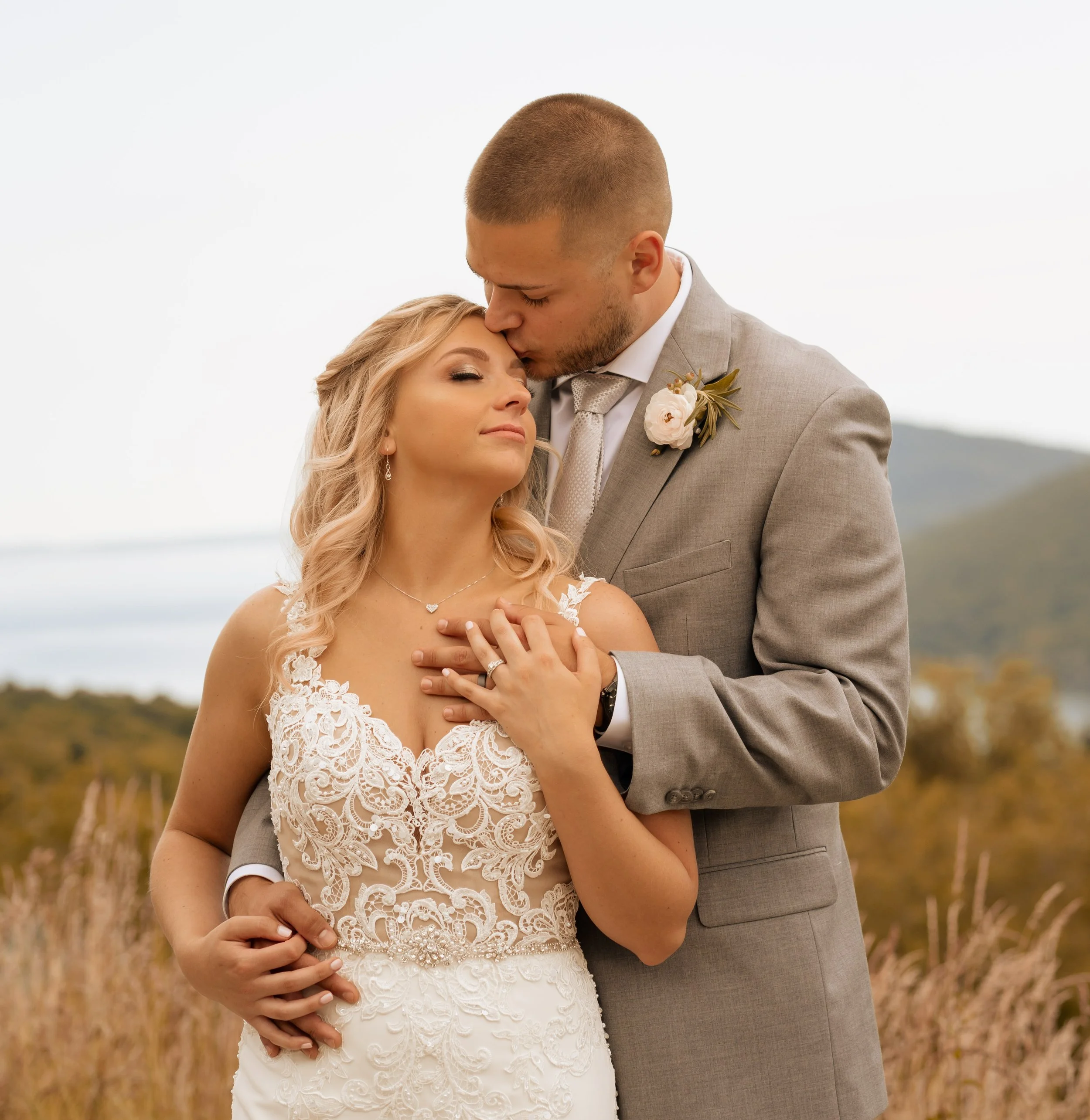 A bride and groom in wedding attire sharing an intimate moment outdoors, with the groom kissing the bride's forehead and her eyes closed, in a natural landscape setting.