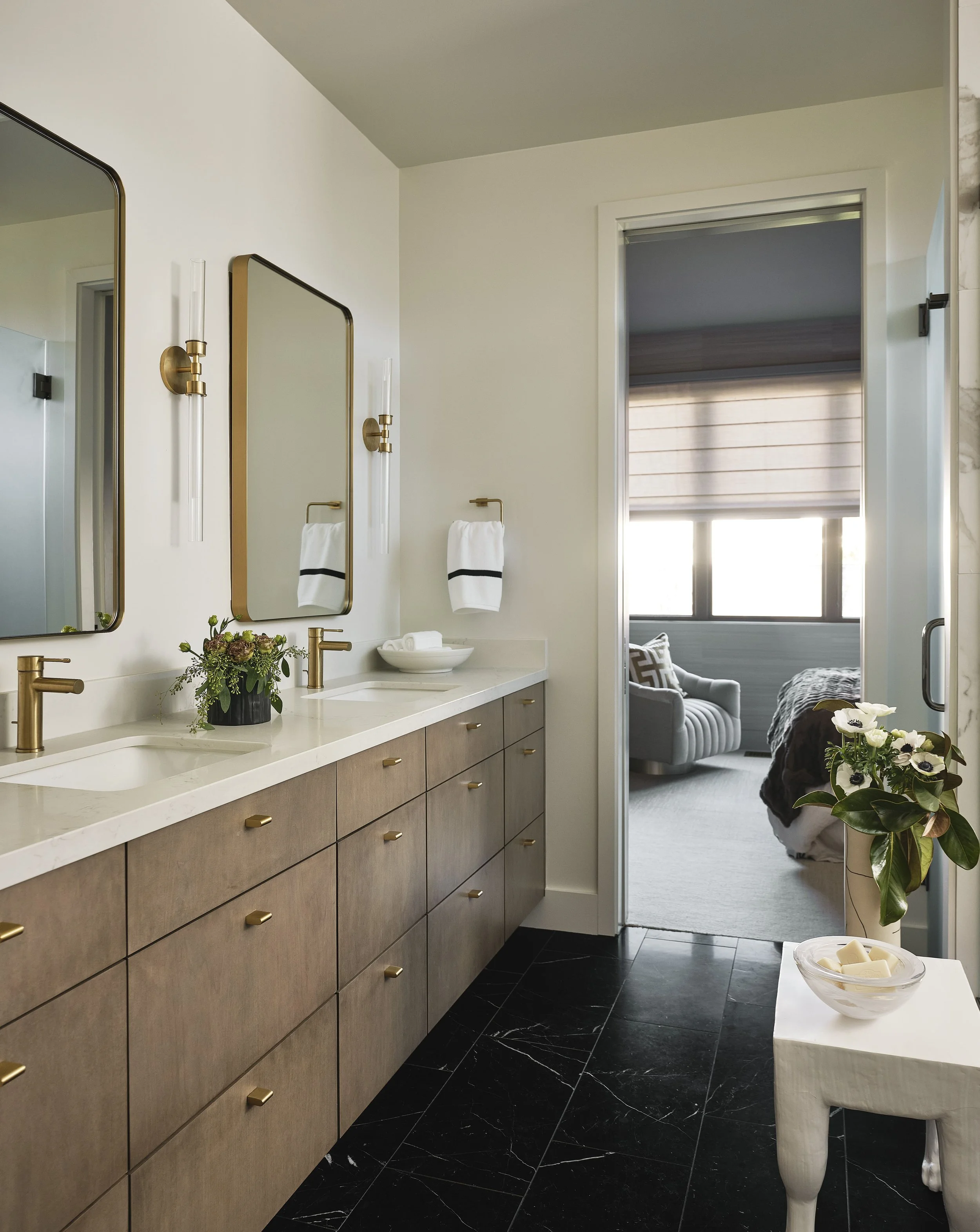 Modern bathroom with double vanity, marble countertop, gold fixtures, large mirrors, and towels, leading to a bedroom with a armchair and window with blinds.