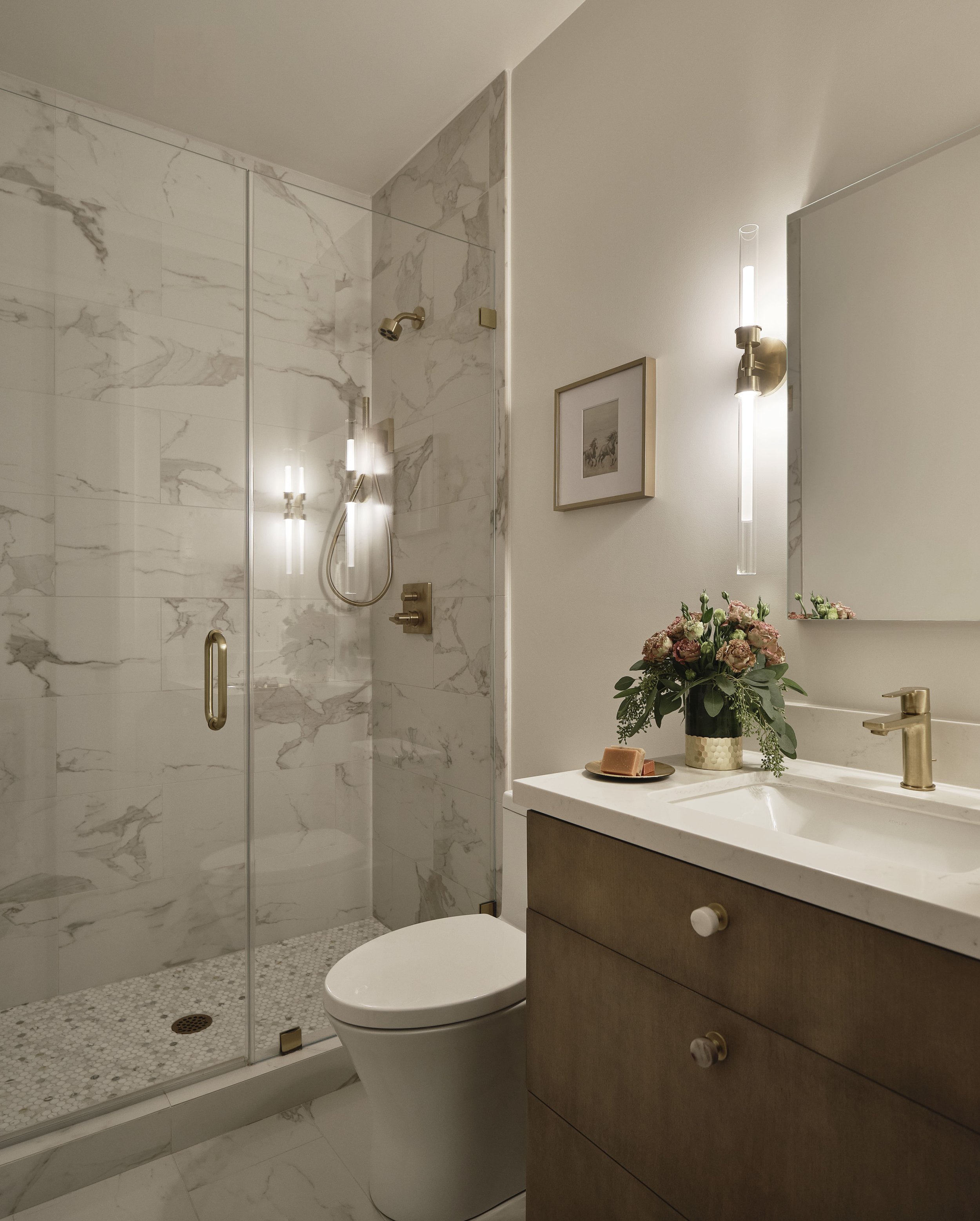 Modern bathroom with marble shower, white toilet, vanity with flower arrangement, and wall-mounted lighting.