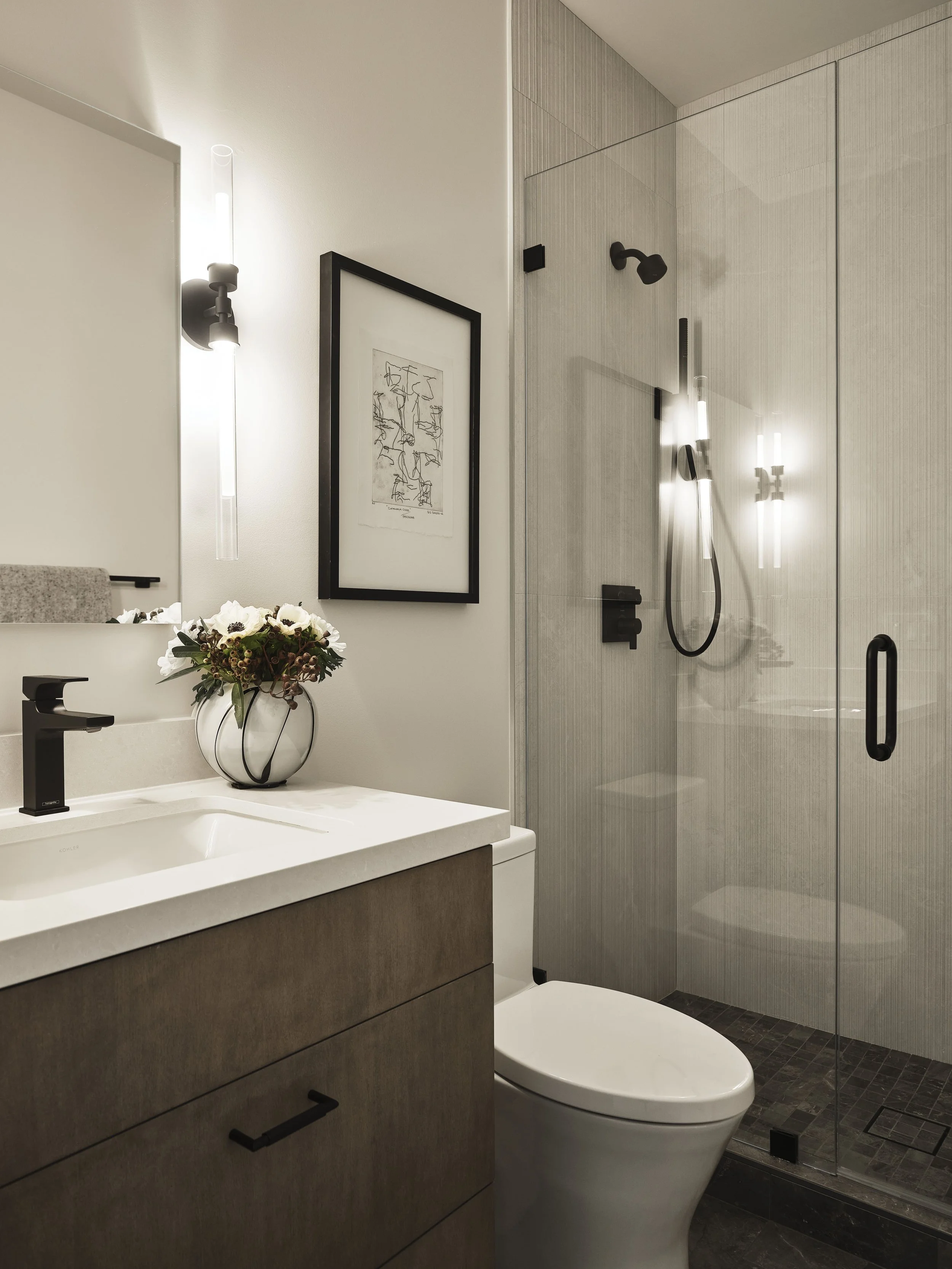 Modern bathroom with white sink, brown vanity, black faucet, wall art, flower vase, toilet, and glass-enclosed shower with black fixtures.