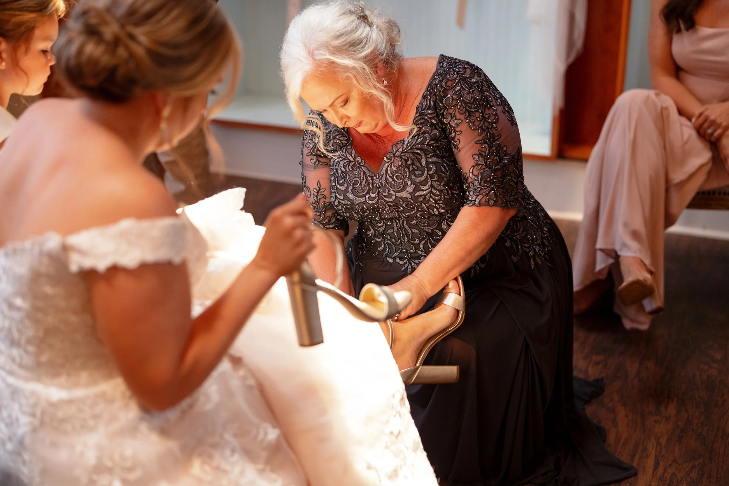 An elderly woman is helping a bride put on her shoe during a wedding preparation in a room.