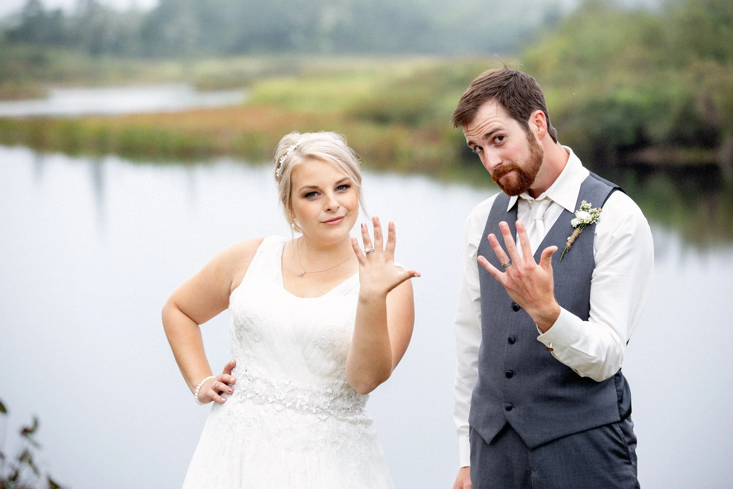 A bride and groom showing their wedding rings outdoors near a lake or river, with trees and water in the background.