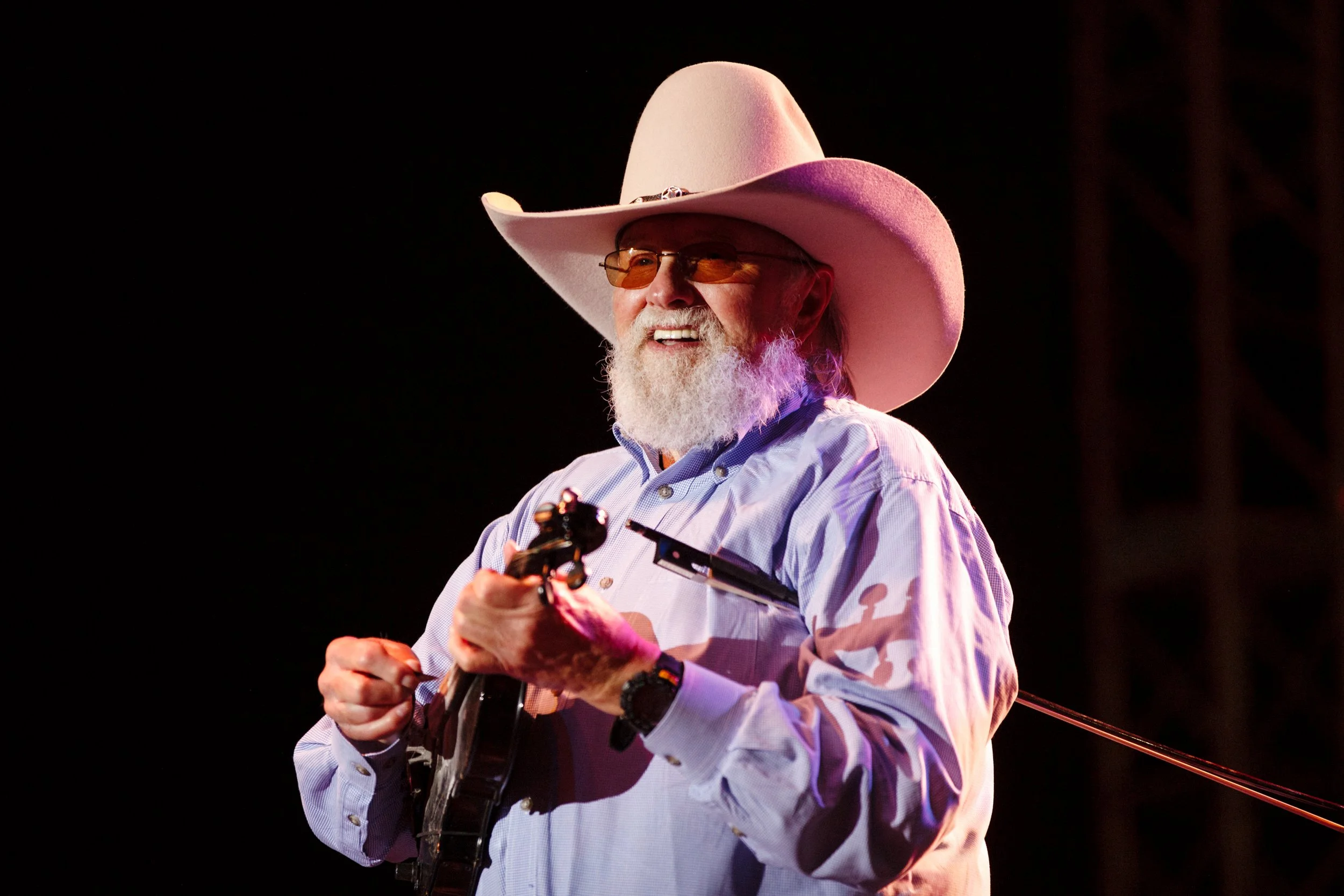 An older man with a white beard wearing a cowboy hat, sunglasses, and a light blue shirt, smiling while playing an acoustic guitar on stage.