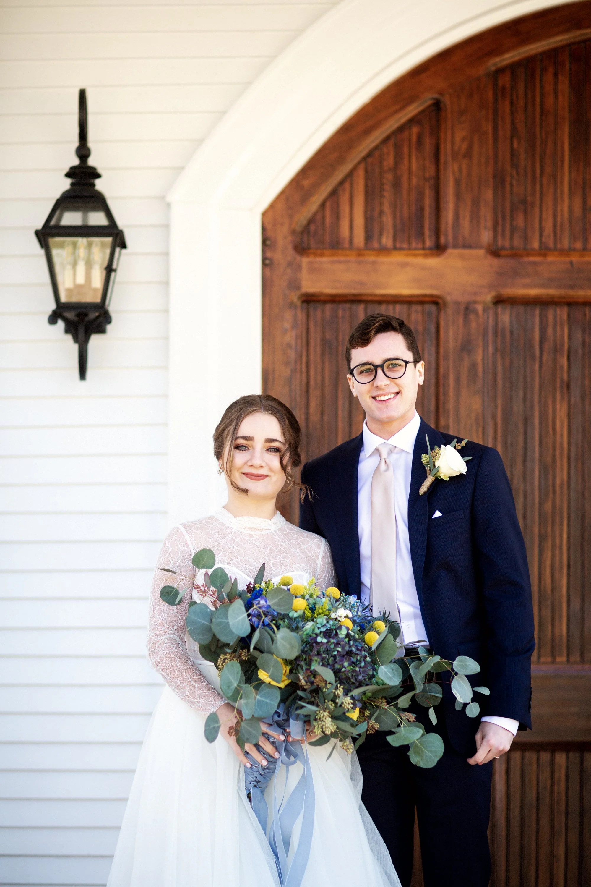 A joyful bride and groom standing in front of a wooden door on their wedding day. The bride holds a large bouquet of green, yellow, and blue flowers, and both are dressed in formal wedding attire. The bride wears a white lace dress; the groom wears a