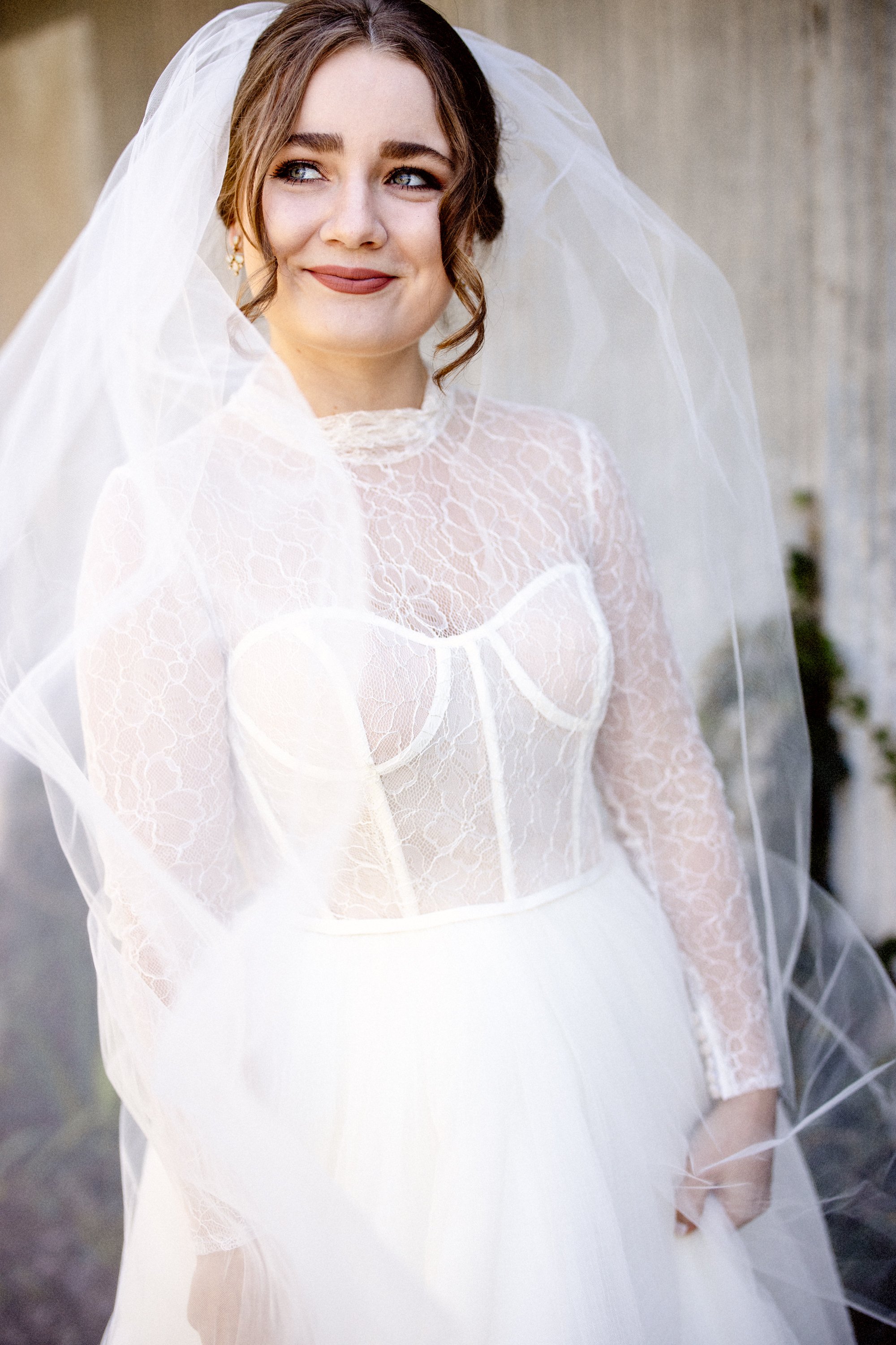 A woman in a wedding dress with lace and a veil, smiling softly and looking slightly away.