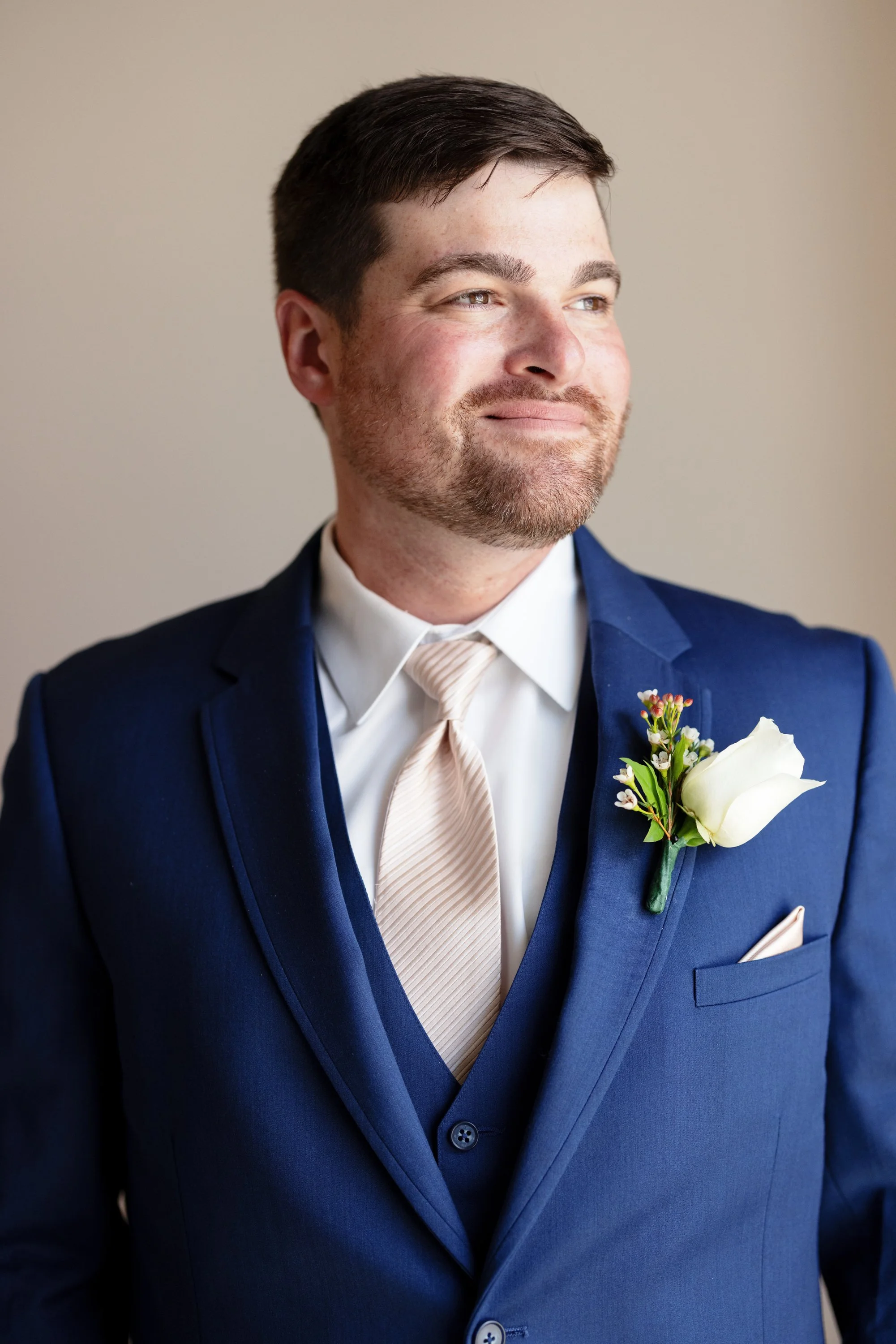 A man in a blue suit with a cream-colored tie and a white boutonniere on his lapel is smiling and looking to the side.