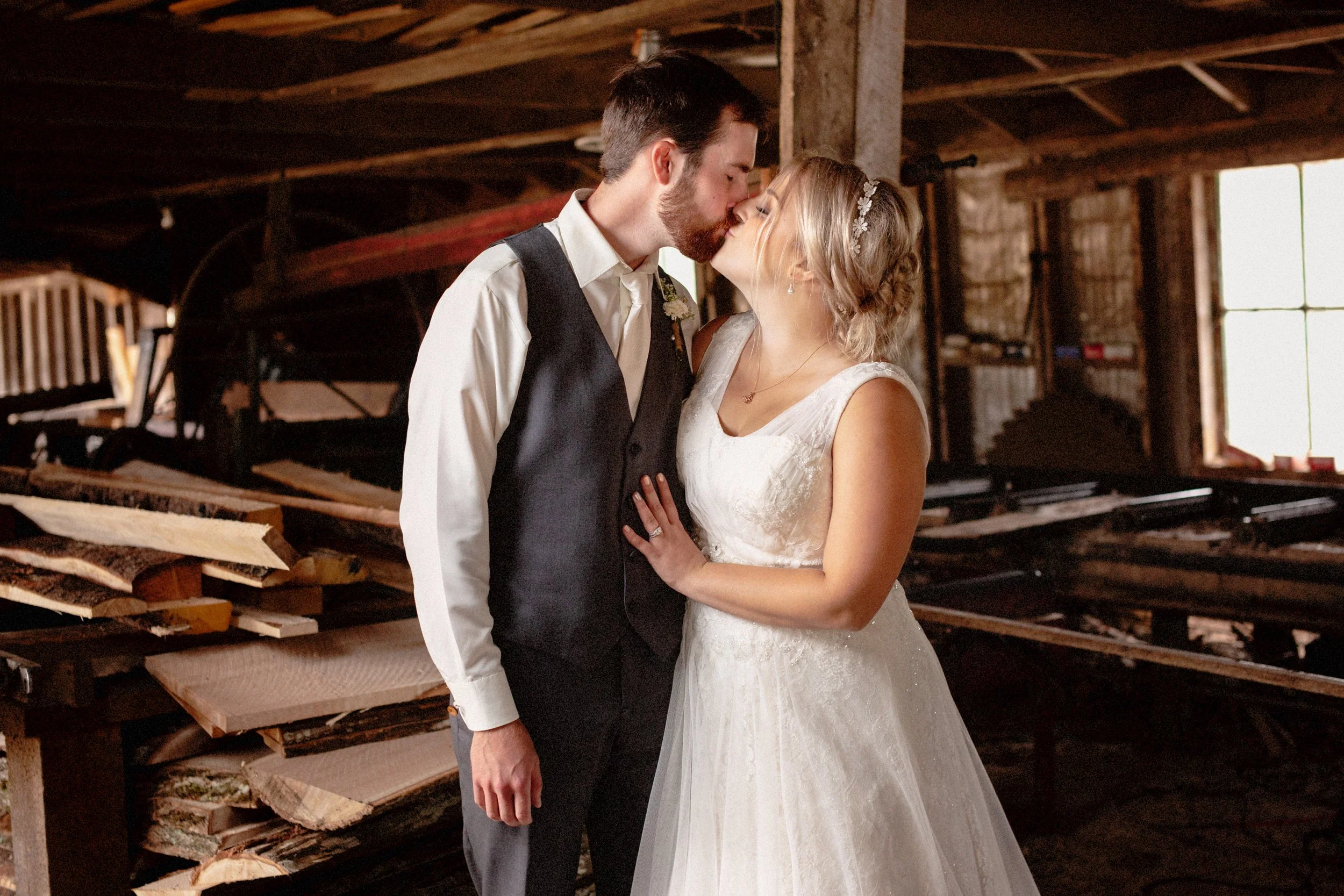 A newlywed couple sharing a kiss in a rustic barn, surrounded by wood logs and machinery, with a large window letting in natural light.