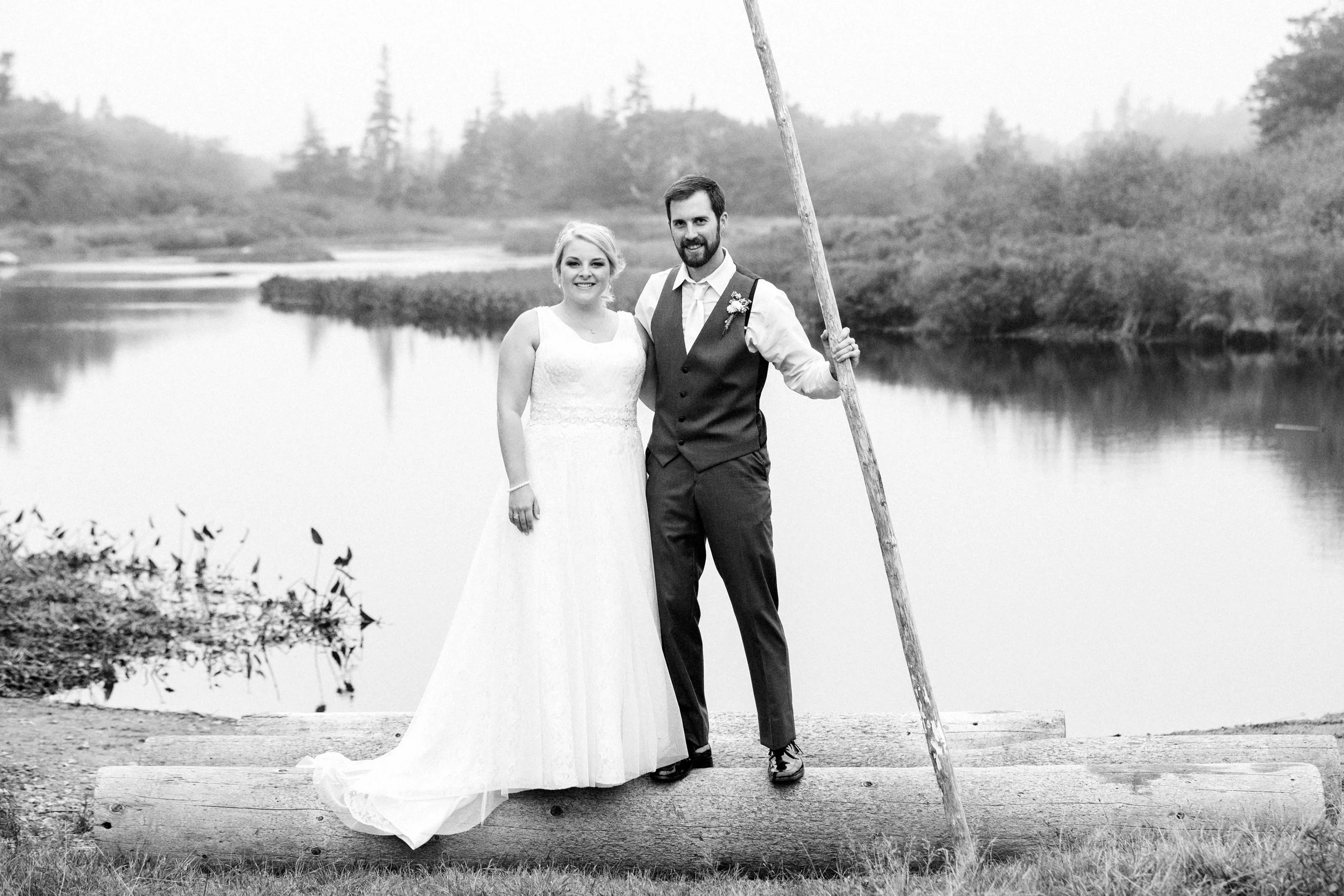 Black and white photo of a bride in a long white dress and groom in a vest and pants standing on a log by a river, holding a long stick, with trees and water in the background.