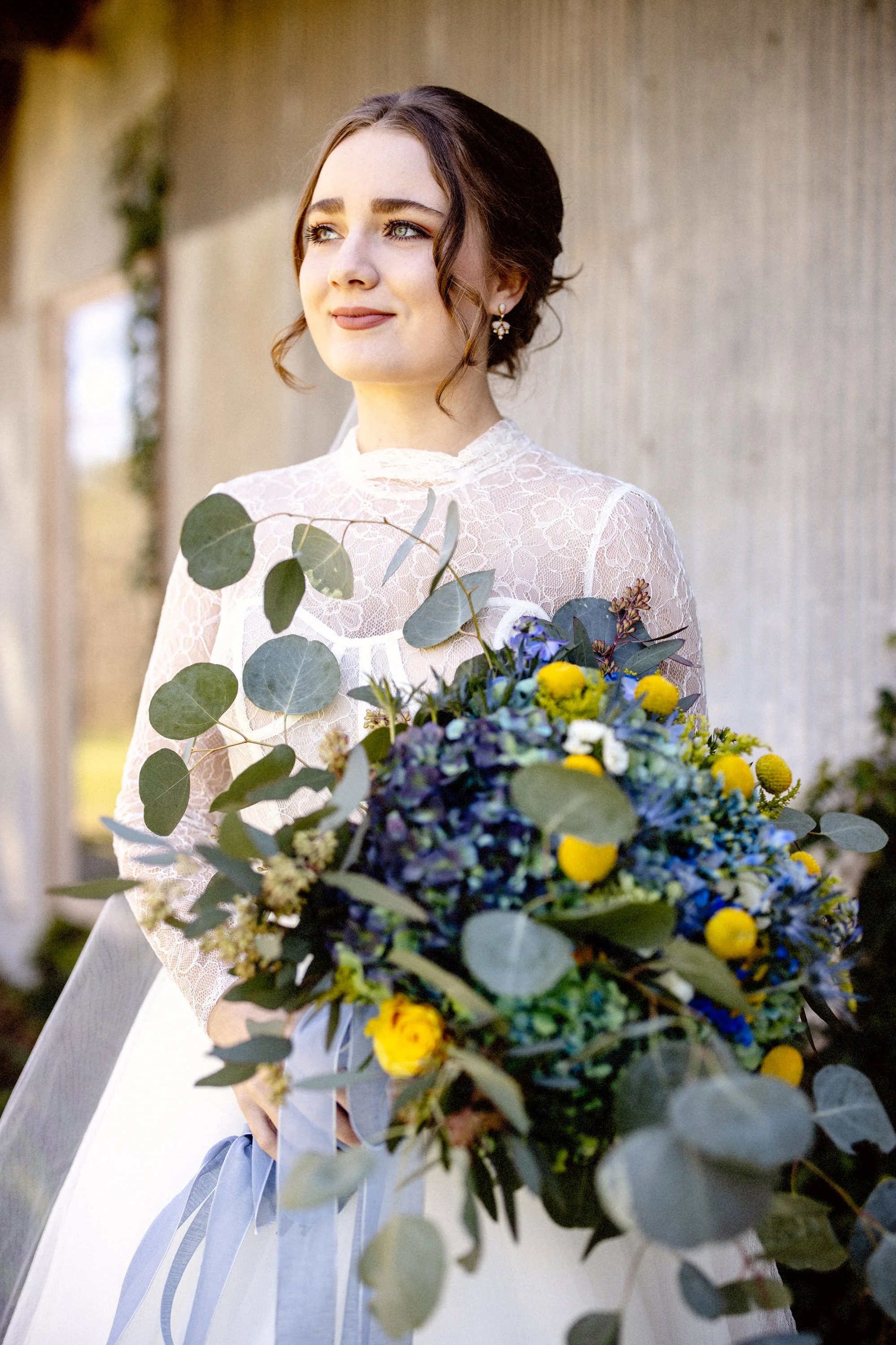 A woman in a white lace top holding a large bouquet of blue, yellow, and green flowers, standing outdoors near a textured wall.