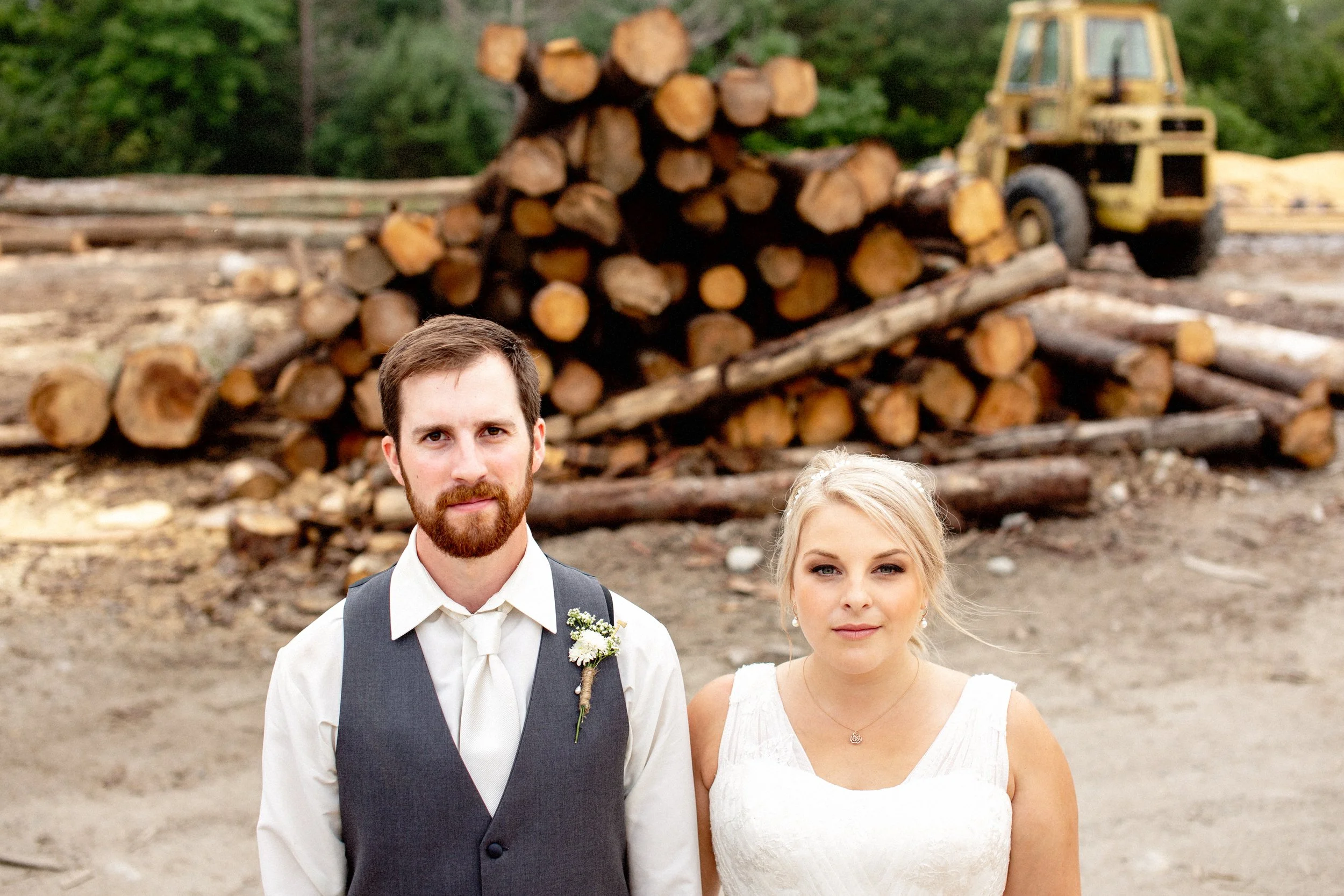 A bride and groom standing outdoors in front of a large pile of logs and construction equipment, with trees in the background.