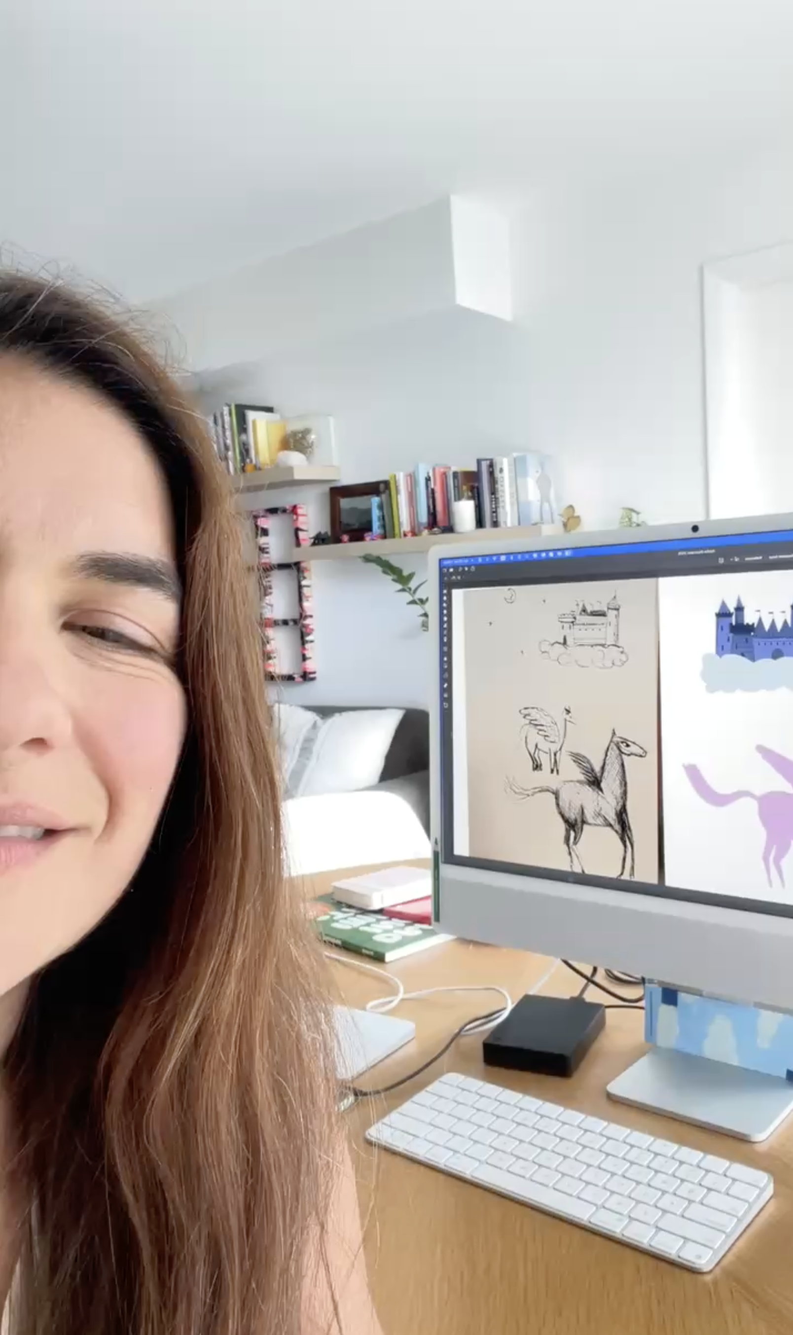 A woman with long reddish-brown hair smiling in a home office, with a computer monitor displaying sketches of a horse, a castle, and a unicorn, on a wooden desk with a keyboard, mouse, and notebooks. In the background, there are white shelves filled 