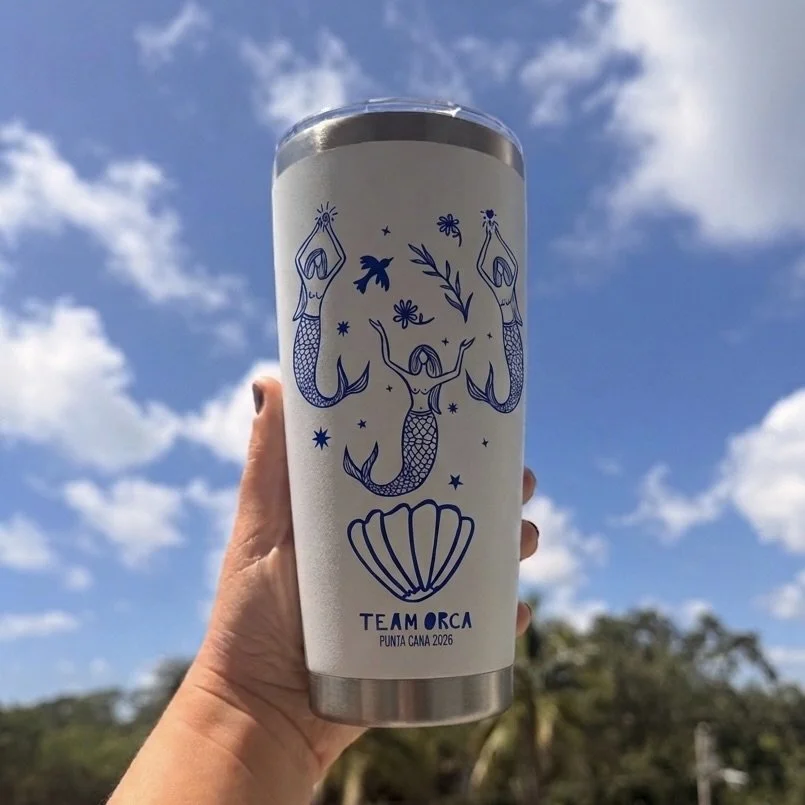 A tall stainless steel tumbler with a white background featuring blue line art illustrations of mermaids, seashells, and stars, held up against a bright sky with clouds and trees in the background.