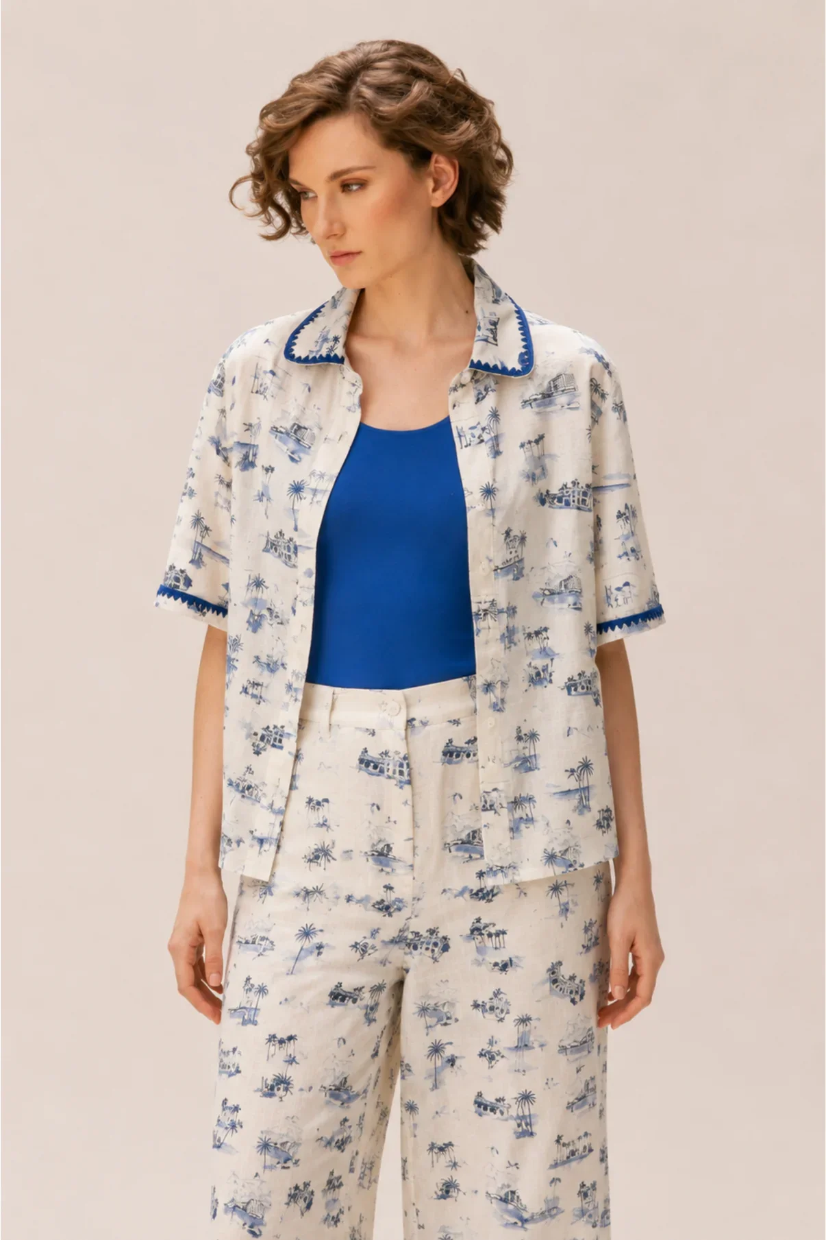 Woman with curly short hair wearing a blue shirt, patterned short-sleeve shirt, and matching pants with a tropical print.
