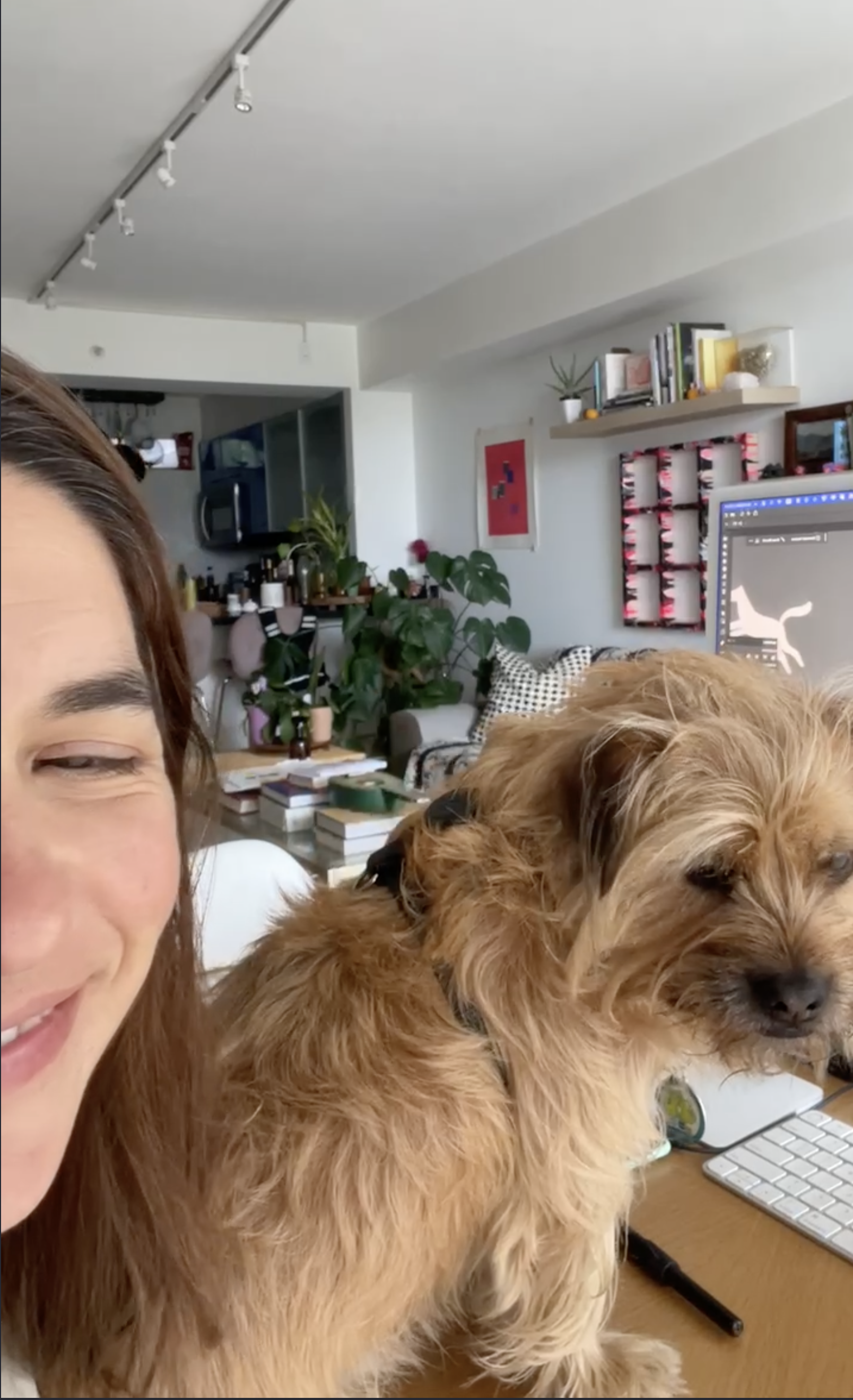 A woman and a fluffy brown dog in a home office or living room. The woman is smiling, and the dog is sitting on a desk with a computer keyboard and a stylus. The room has plants, books, and decorative items, with a bookshelf on the wall and a framed 