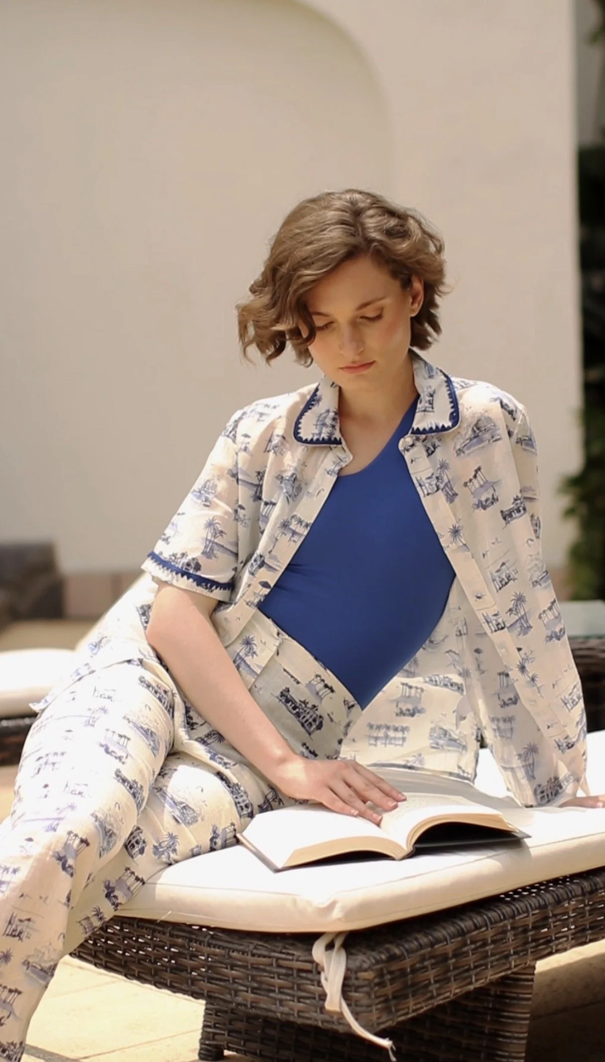 A woman with short wavy brown hair wearing a coordinated pajama set with beach and palm tree pattern, sitting on a wicker outdoor lounge chair with a cushion, reading a book outdoors.