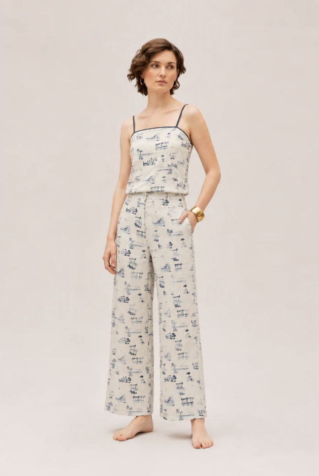 A woman standing barefoot against a plain neutral background, wearing a matching sleeveless top and wide-leg pants set with a blue and beige toile pattern, and accessorized with a gold bracelet.