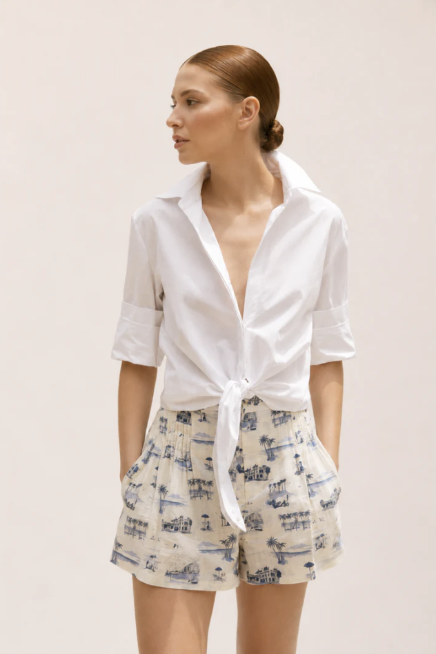 A woman with reddish hair in a low bun wearing a white tied-up button-up shirt and printed shorts with beach scenes.