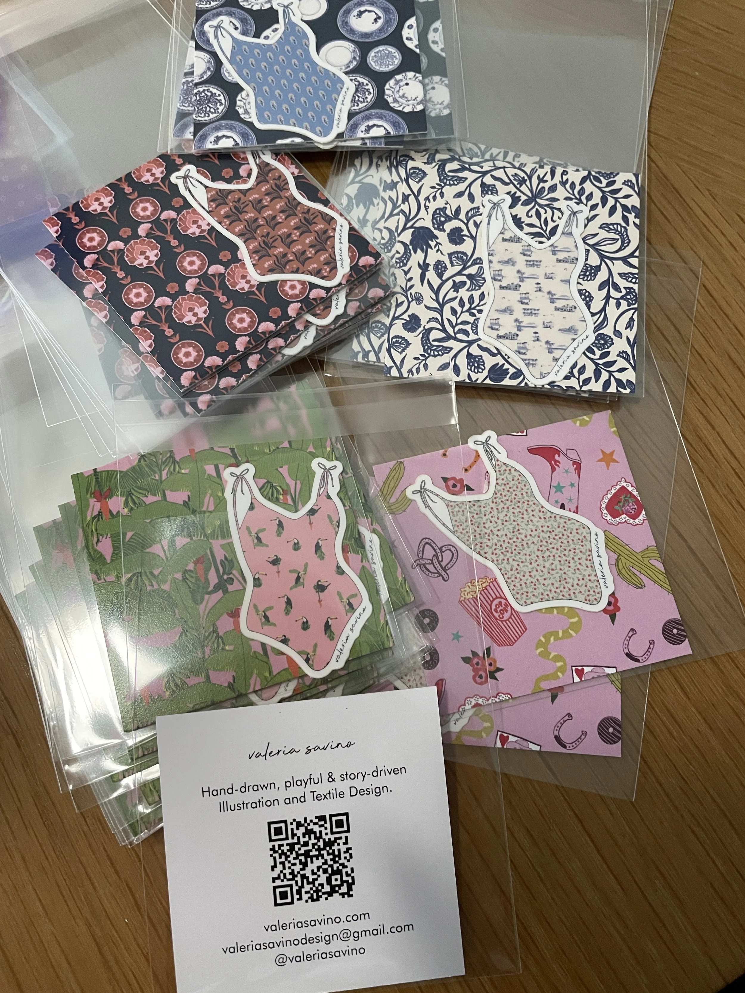 Sets of colorful illustrated paper bags with various patterns and a white card with contact information, all in a transparent plastic folder. The paper bags have playful, story-driven textile designs.