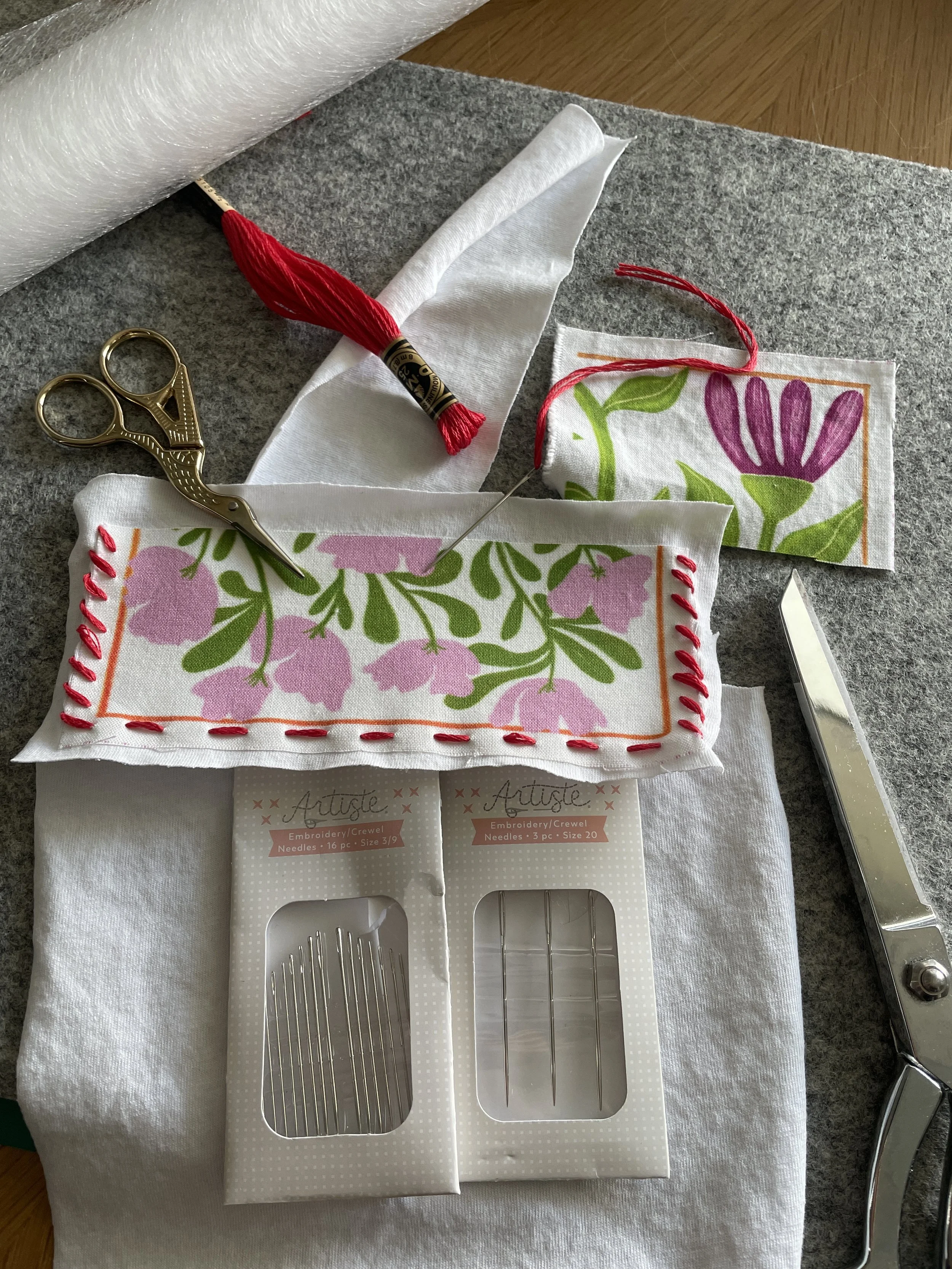 Embroidery tools including a pair of gold scissors, a needle threader, two packages of embroidery needles, a needle, a piece of fabric with pink floral and green leaf design, and a metallic tool, all on a gray surface.
