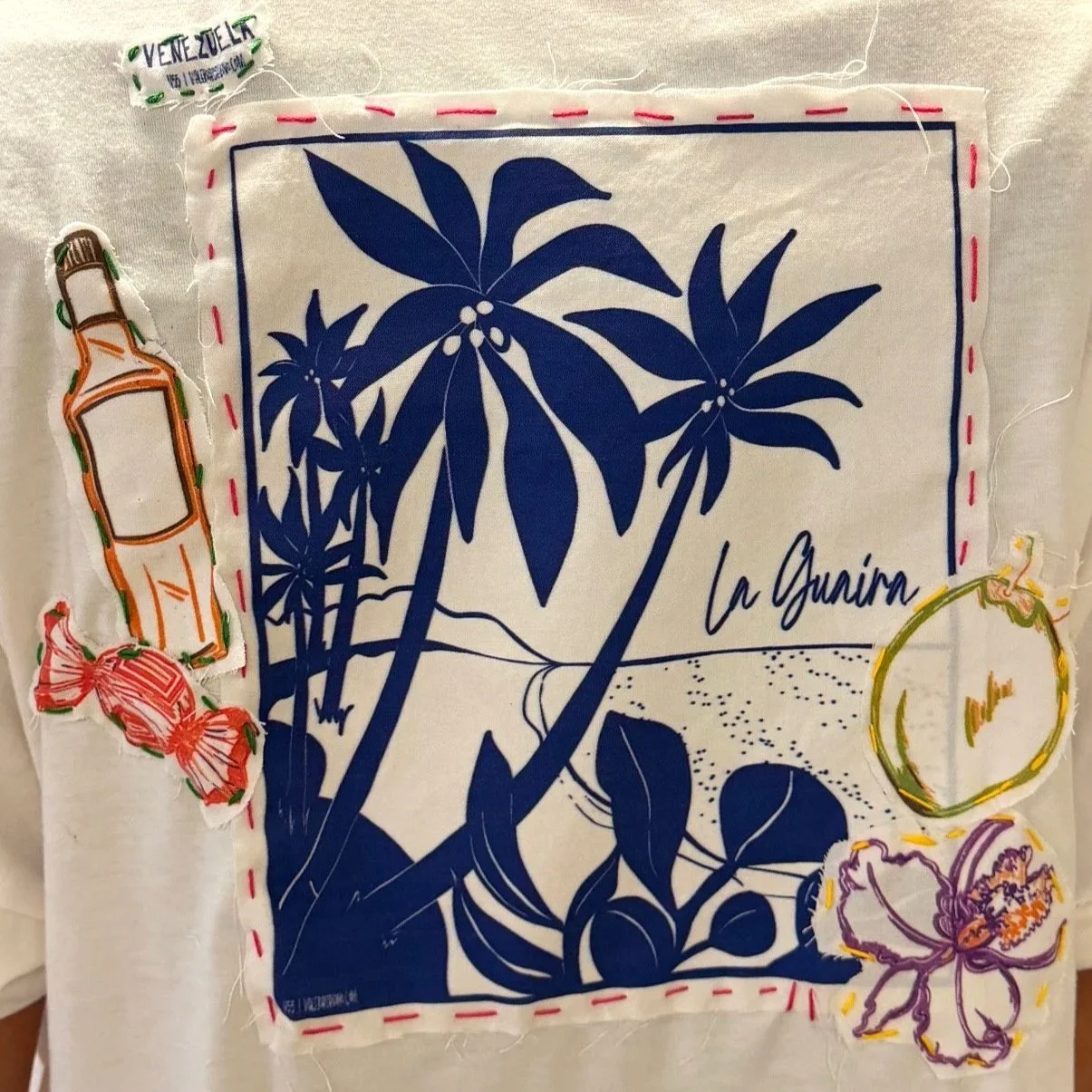 Fabric patch featuring a blue silhouette of a beach scene with palm trees, mountains, and sea, with the text "La Guaira." Surrounding the patch are stitched and embroidered illustrations of a bottle, a face, a butterfly, a round object, and candies, all on a white background.