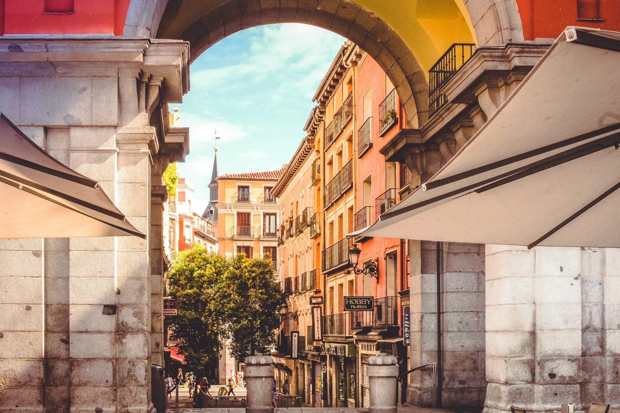 Arched stone entrance to a European street with colorful old buildings, balconies, and people walking; canopies frame the sides.