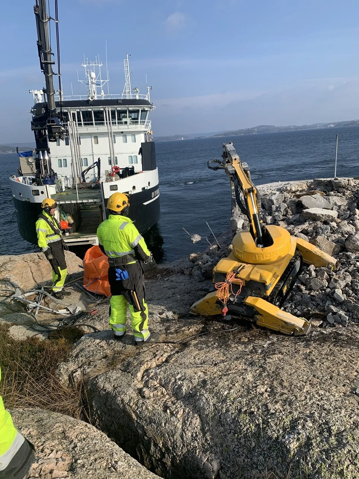 Brokk rivemaskin på kystskjær med fartøy og mannskap i bakgrunnen