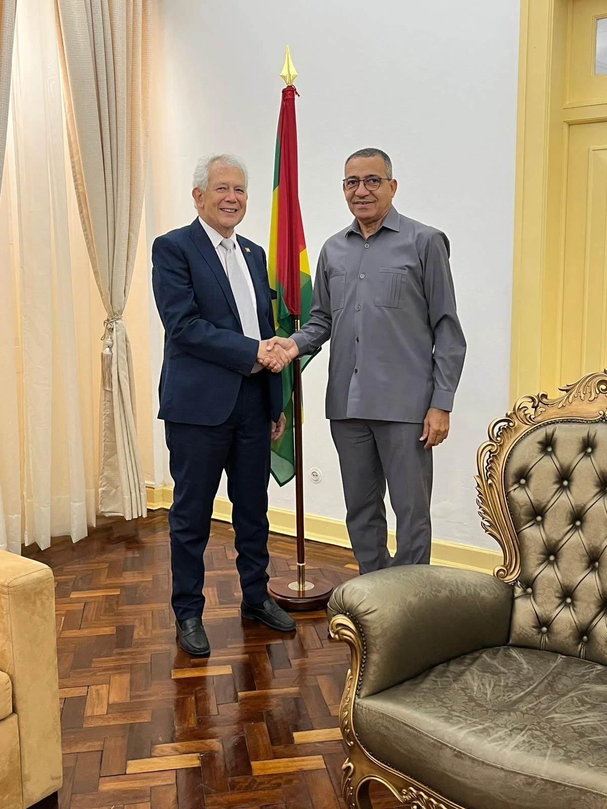 Un homme en costume avec une cravate rouge et des lunettes, assis devant un micro, avec un fond flou de couleur verte.