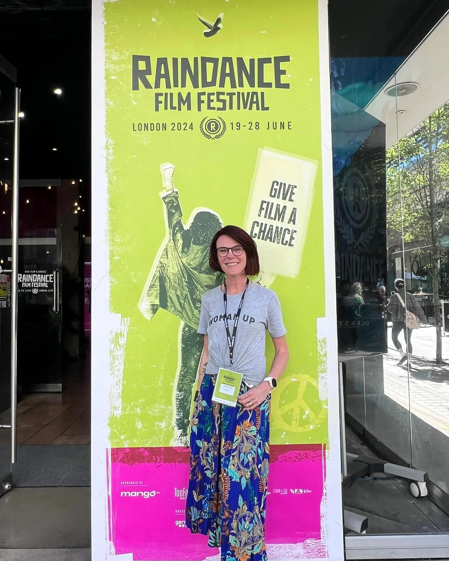 Johanna Courtney, standing in front of a large promotional poster for the Raindance Film Festival in London, 2024, smiling, wearing glasses, a grey T-shirt with the words 'WOMAN UP' printed on it, and a blue floral skirt. 