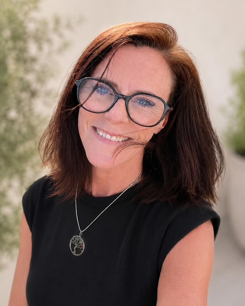Johanna Courtney, smiling, outdoors, wearing glasses, a black top, and a necklace with a tree of life pendant, with a blurred background of greenery.