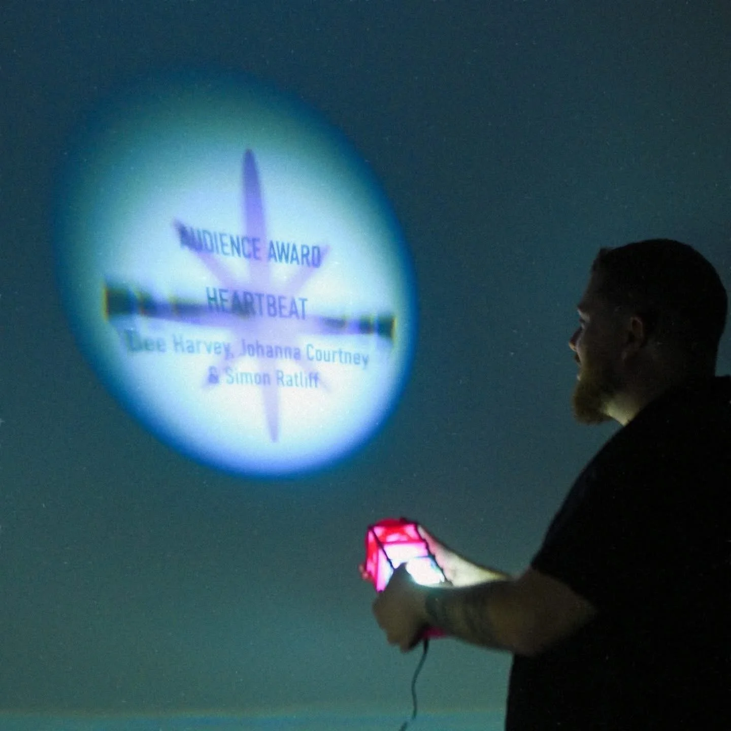 Ondra Moravec, announcing the winner of the Audience Award with a projection on a dark wall for the film 'Heartbeat,' with the names Dee Harvey, Johanna Courtney, and Simon Rattif., ART*VR 2024.