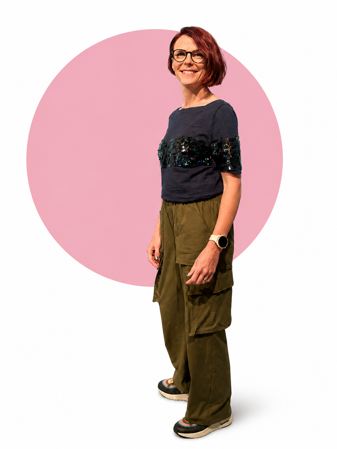 Johanna Courtney, smiling, with short red hair, glasses, wearing a dark top with sequin embellishments on sleeves, khaki cargo pants, Nike trainers, and a Garmin watch, standing in front of a pink circle on a white background.