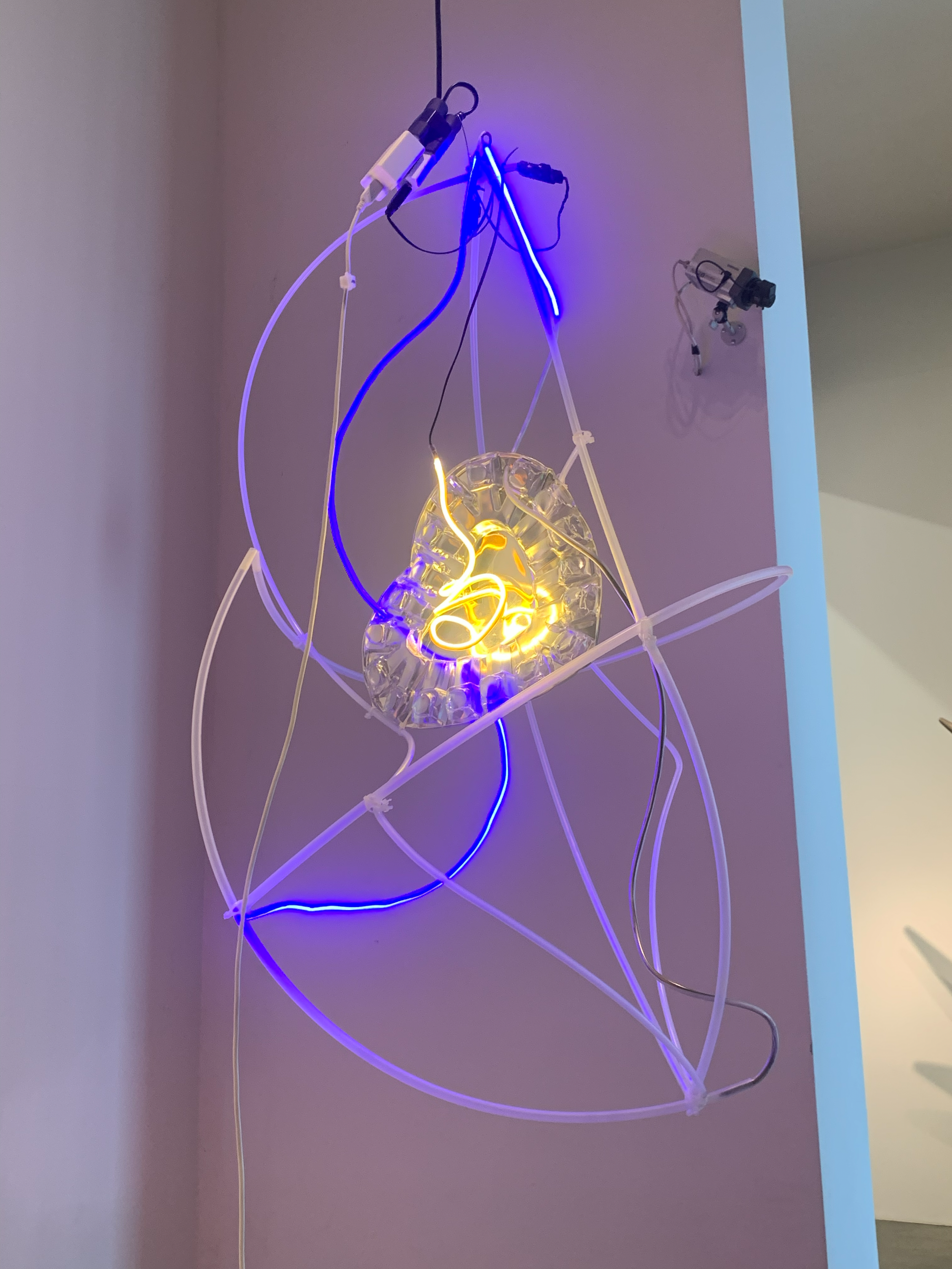 Abstract interpretation from the animation in Heartbeat - wall art with a three-dimensional clear acrylic disc and illuminated blue, yellow, and white neon lights, mounted on a corner wall, at the 'Beyond the Glass Ceiling' showcase, ART*VR, 2024.