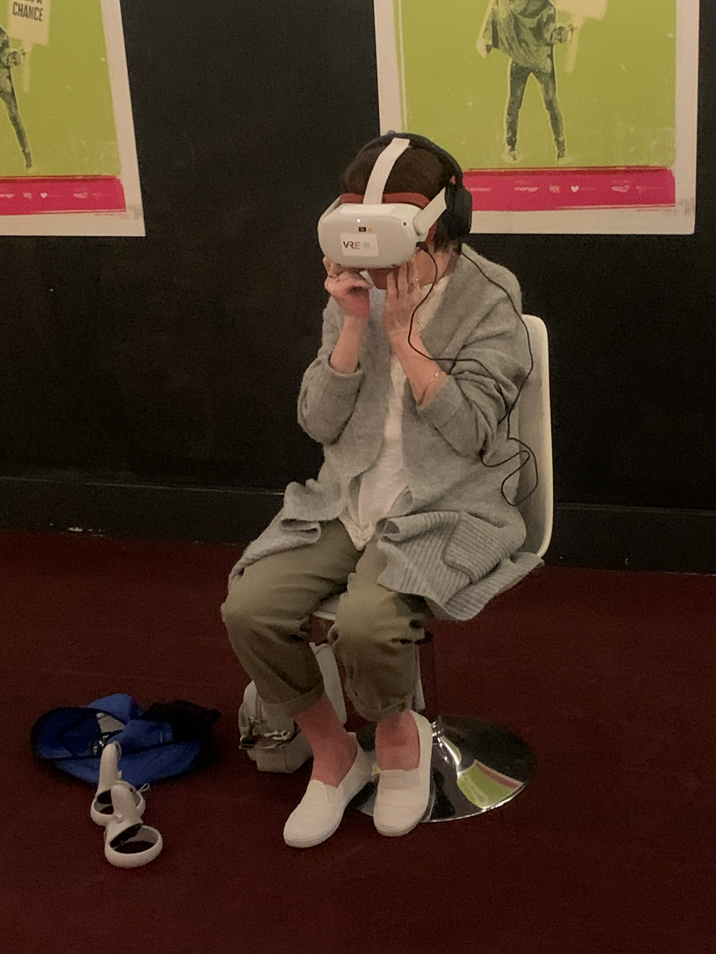 A lady sitting on a stool wearing a virtual reality headset, watching Heartbeat at the Raindance Film Festival, Immersive Summit, 2024. A backpack and belongings are on the floor nearby, in front of Festival posters on a black wall.
