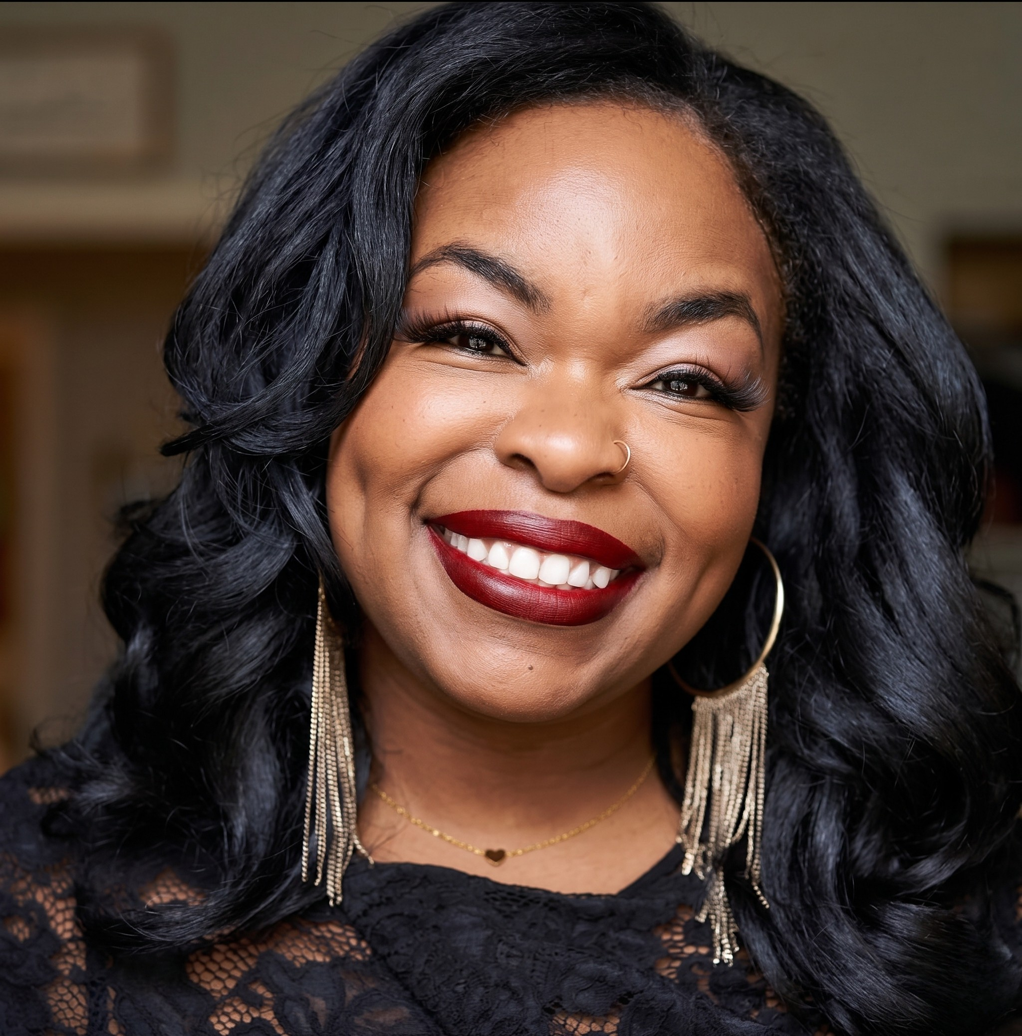 Close-up of a smiling woman with black, wavy hair, wearing red lipstick, gold jewelry, and a black lace top.