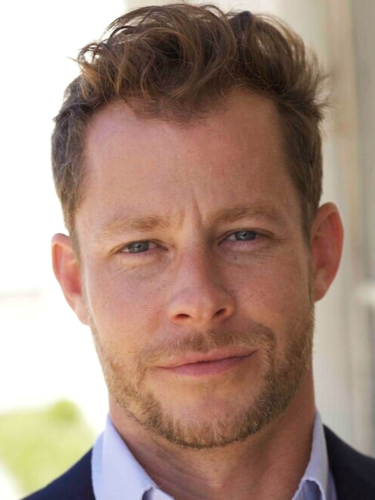 Close-up of a man with light brown hair, blue eyes, and a short beard, wearing a dark blazer and light blue shirt, outdoors in natural light.