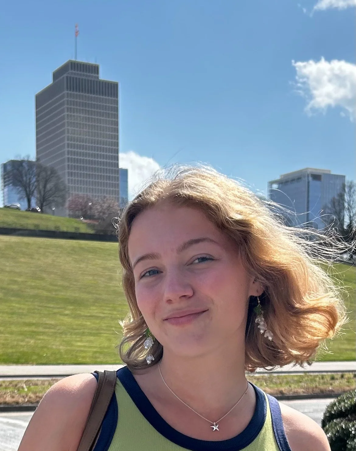 A young woman with short, wavy blond hair outdoors on a sunny day, with a cityscape and blue sky in the background.