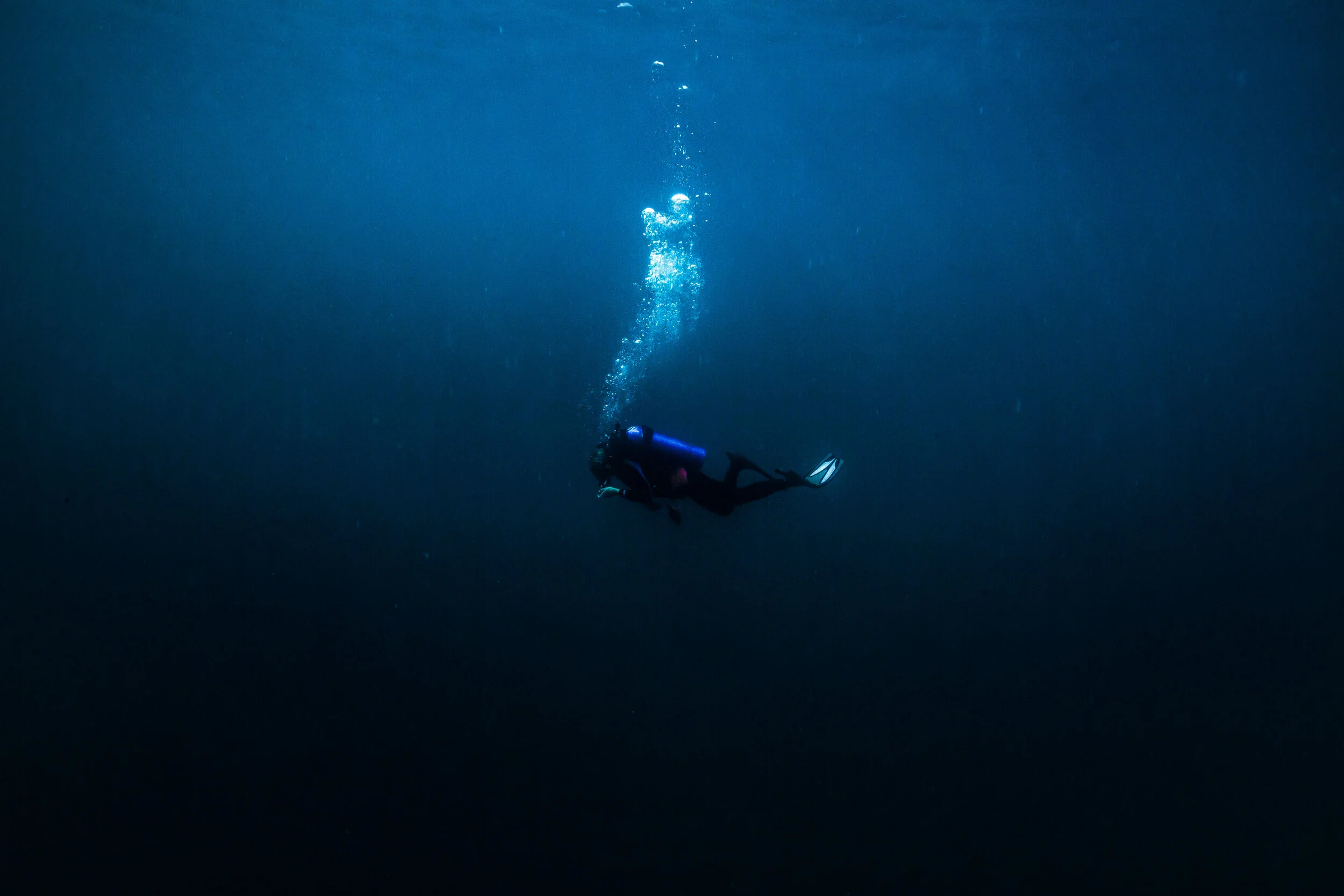 A scuba diver swimming alone in deep blue water, with bubbles rising to the surface.